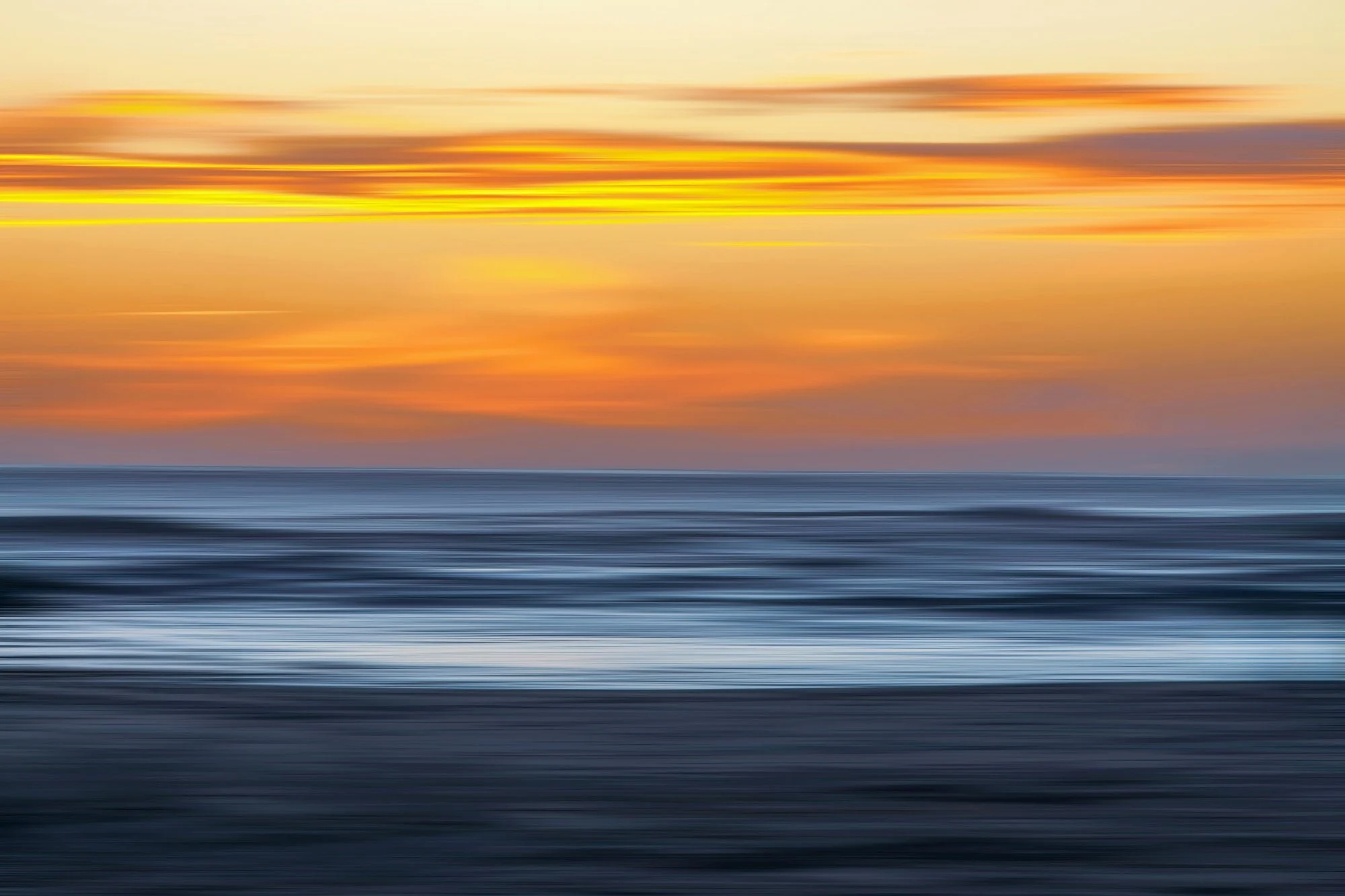 Imagen de un atardecer o amanecer en el mar con líneas de color naranja, amarillo y gris, y un efecto de movimiento que la hace parecer borrosa o como si estuviera en movimiento rápido.