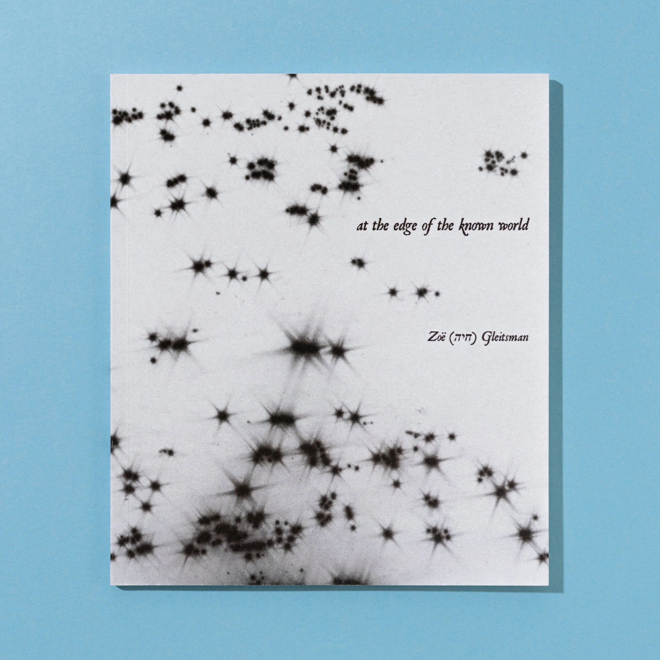 Book titled 'at the edge of the known world' by Zoë Gleitman with a black and white starry sky cover design, placed on a blue background.