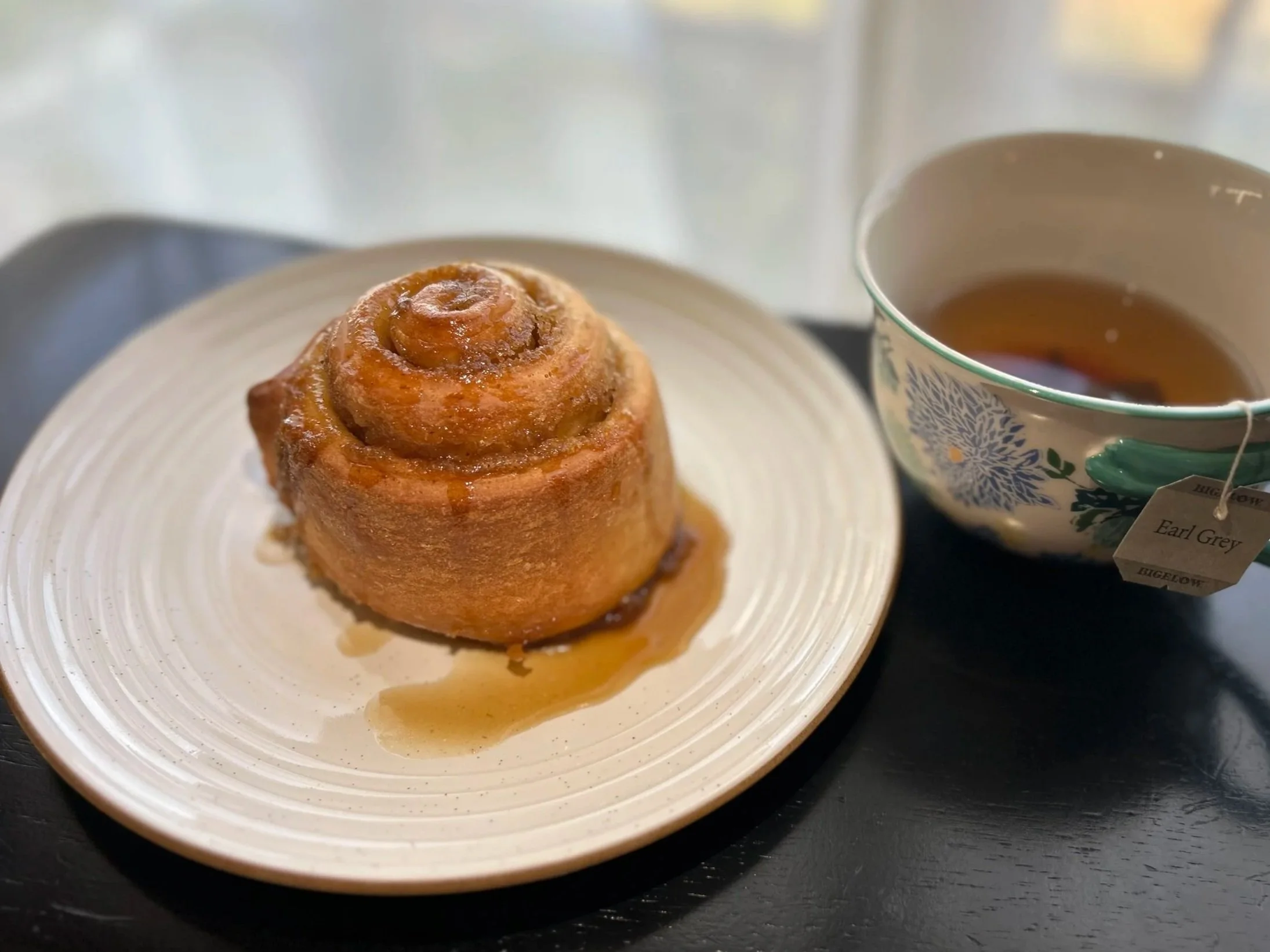 London Fog roll drizzled with glaze on a white plate, next to a cup of tea, labeled Earl Grey, on a black tray.
