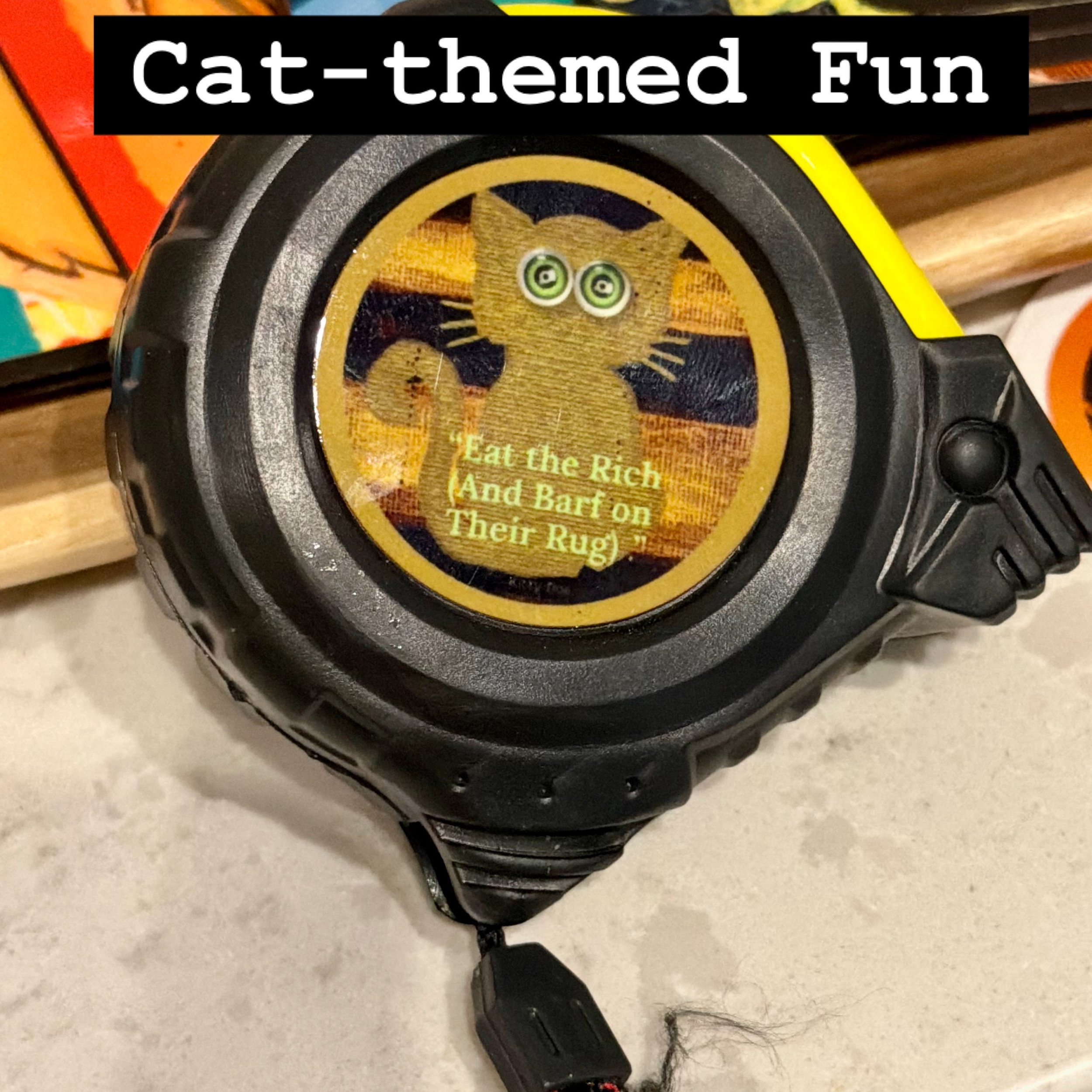 Tape Measure that has a cat with a striped background and says "Eat the rich (and barf on their rug)" in green lettering