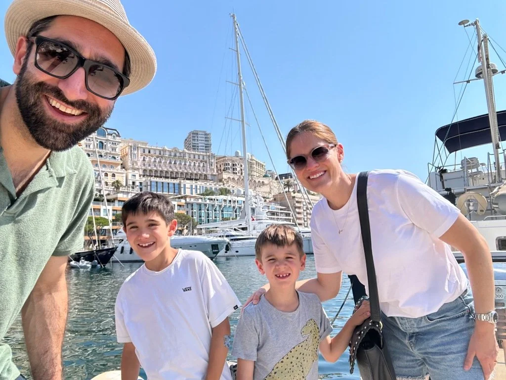 A family of four, including a man, woman, and two boys, smiling at a marina with sailboats and yachts, under a clear blue sky.