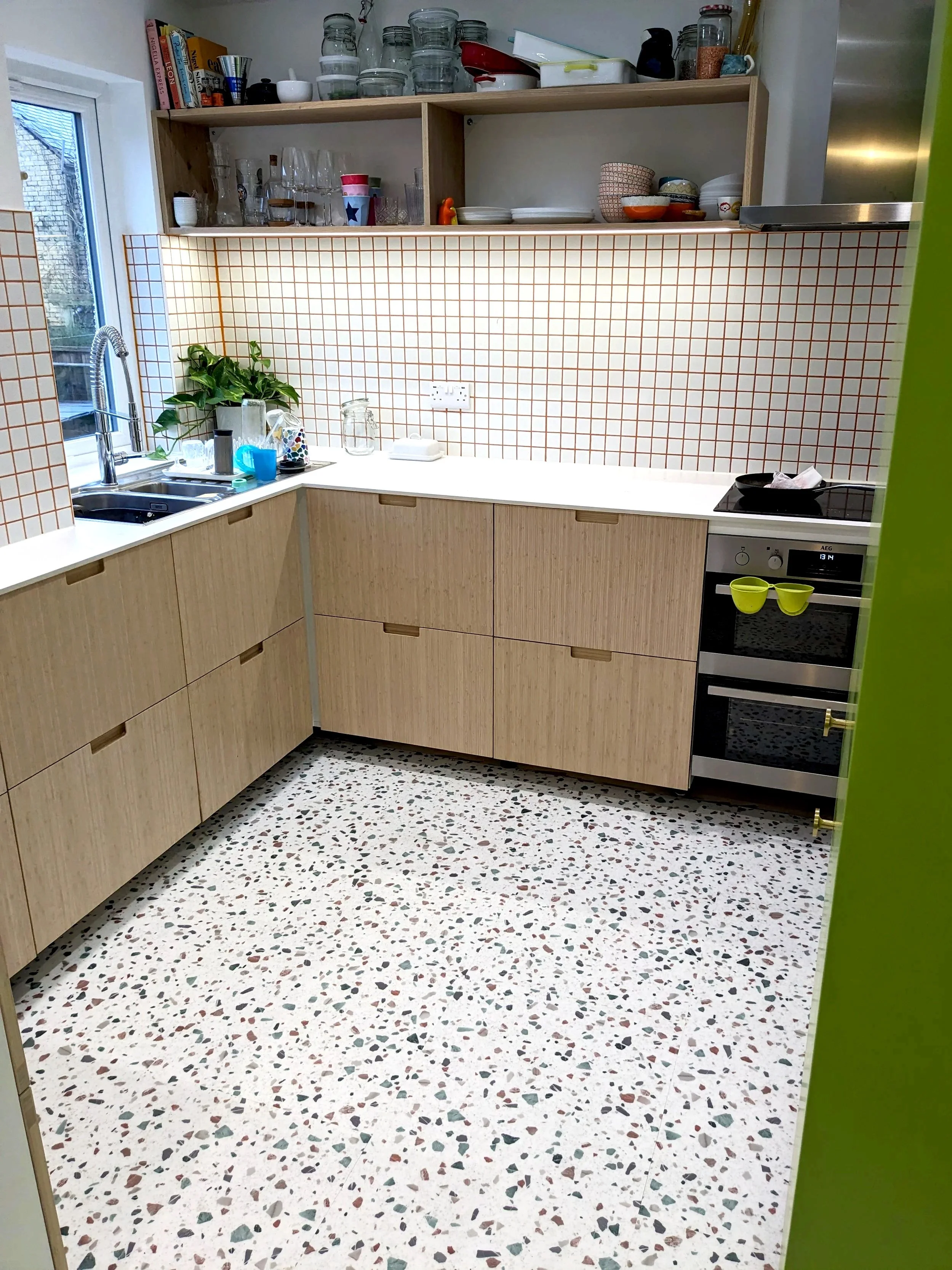 A kitchen with wooden cabinets, a white countertop, a terracotta-tiled backsplash, and a terrazzo floor. There is a sink by a window with some glasses and a plant, and an oven with lime green mugs hanging from it. Open shelves hold glassware, bowls, and kitchen items.