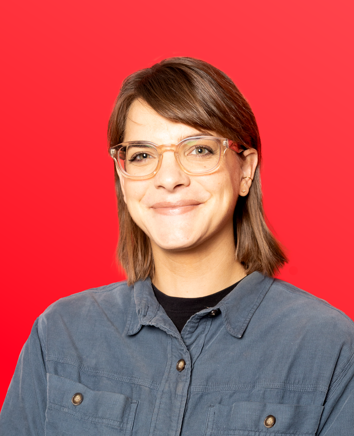 A woman with short brown hair, glasses, and earrings, smiling, wearing a gray button-up shirt against a light blue background.