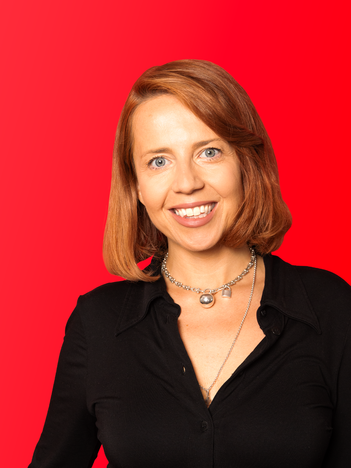 A woman with shoulder-length red hair, wearing a black button-up shirt and silver jewelry, smiling against a red background.