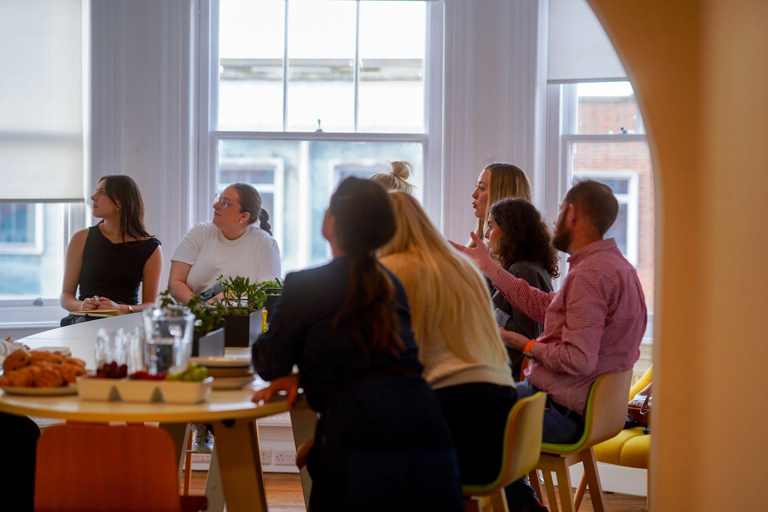 A group of people sitting and standing around a conference table in a brightly lit room, engaging in a discussion.