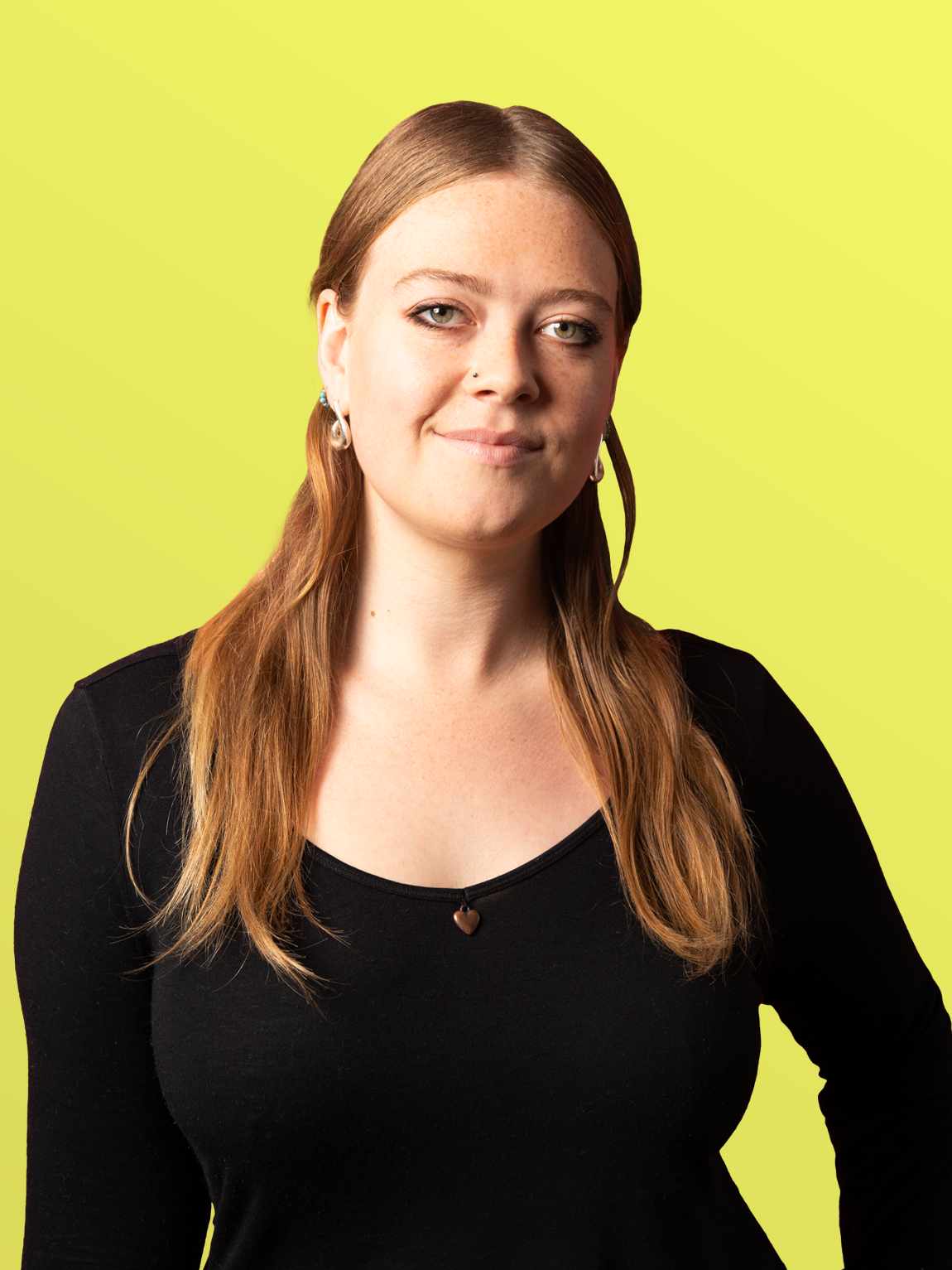 A woman with red hair, light skin, and freckles, wearing a black top and earrings, standing against a yellow background.