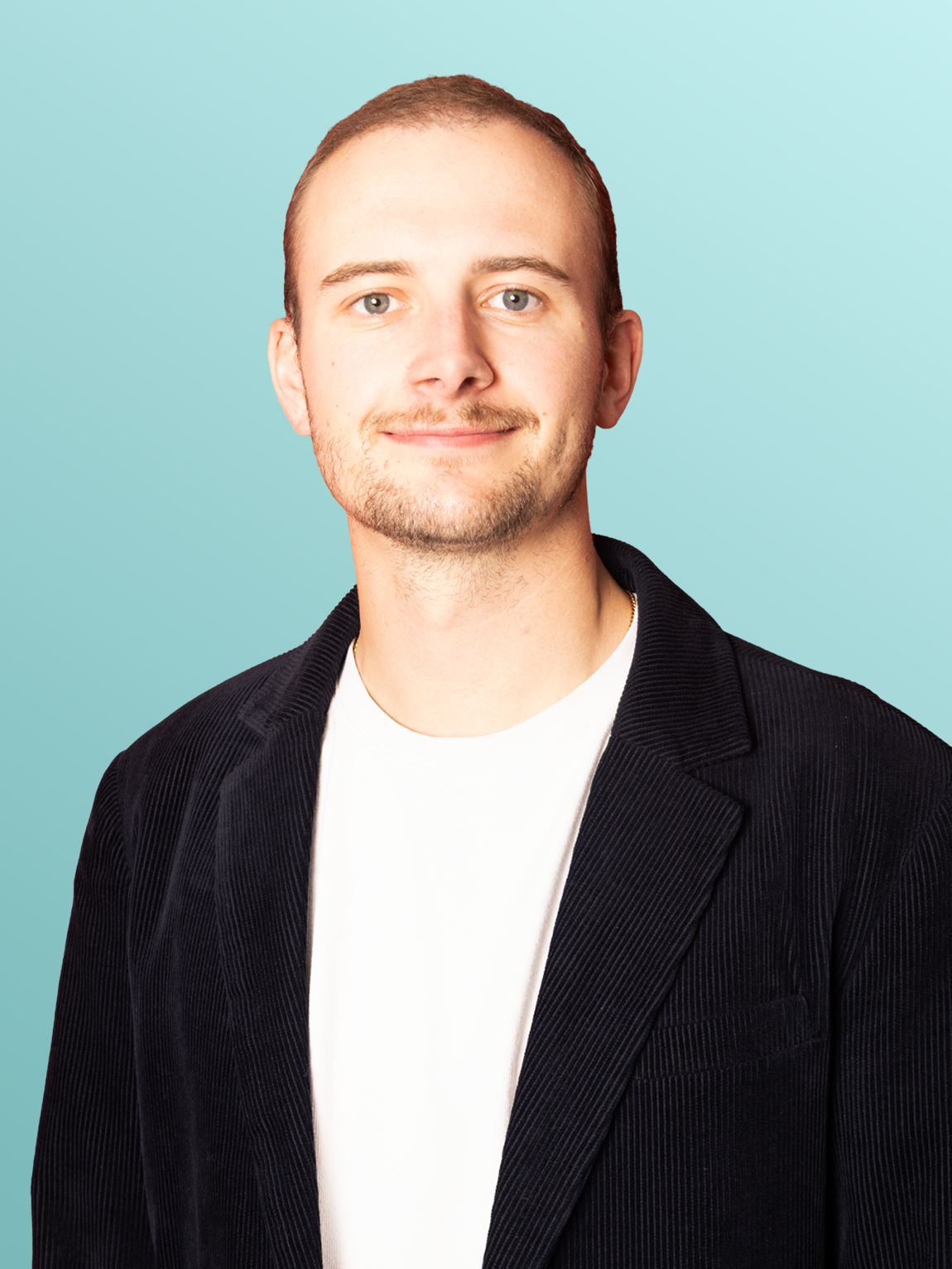 A young man with short brown hair and a light beard, wearing a black blazer over a white t-shirt, smiling against a light blue background.