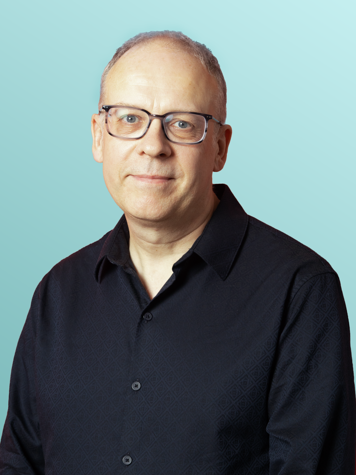 A man wearing glasses and a black button-up shirt stands against a light blue background.