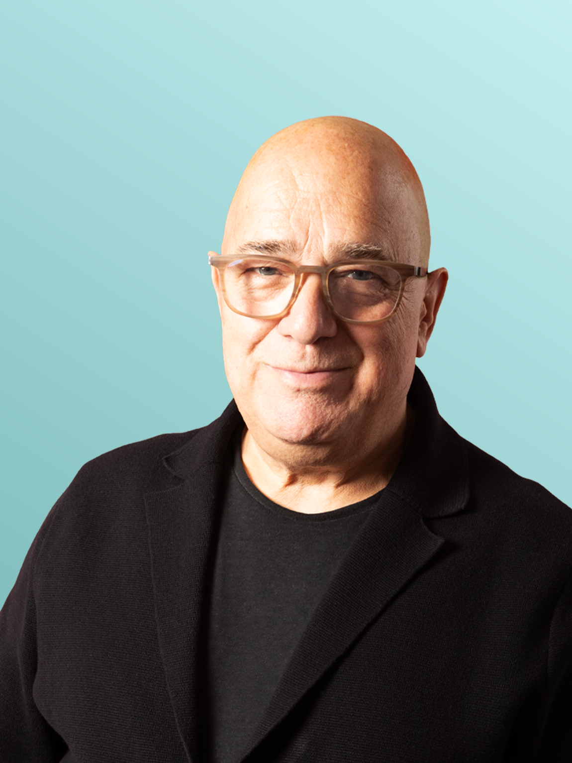 A bald man wearing glasses, a black blazer, and a black shirt, standing against a light yellow background.
