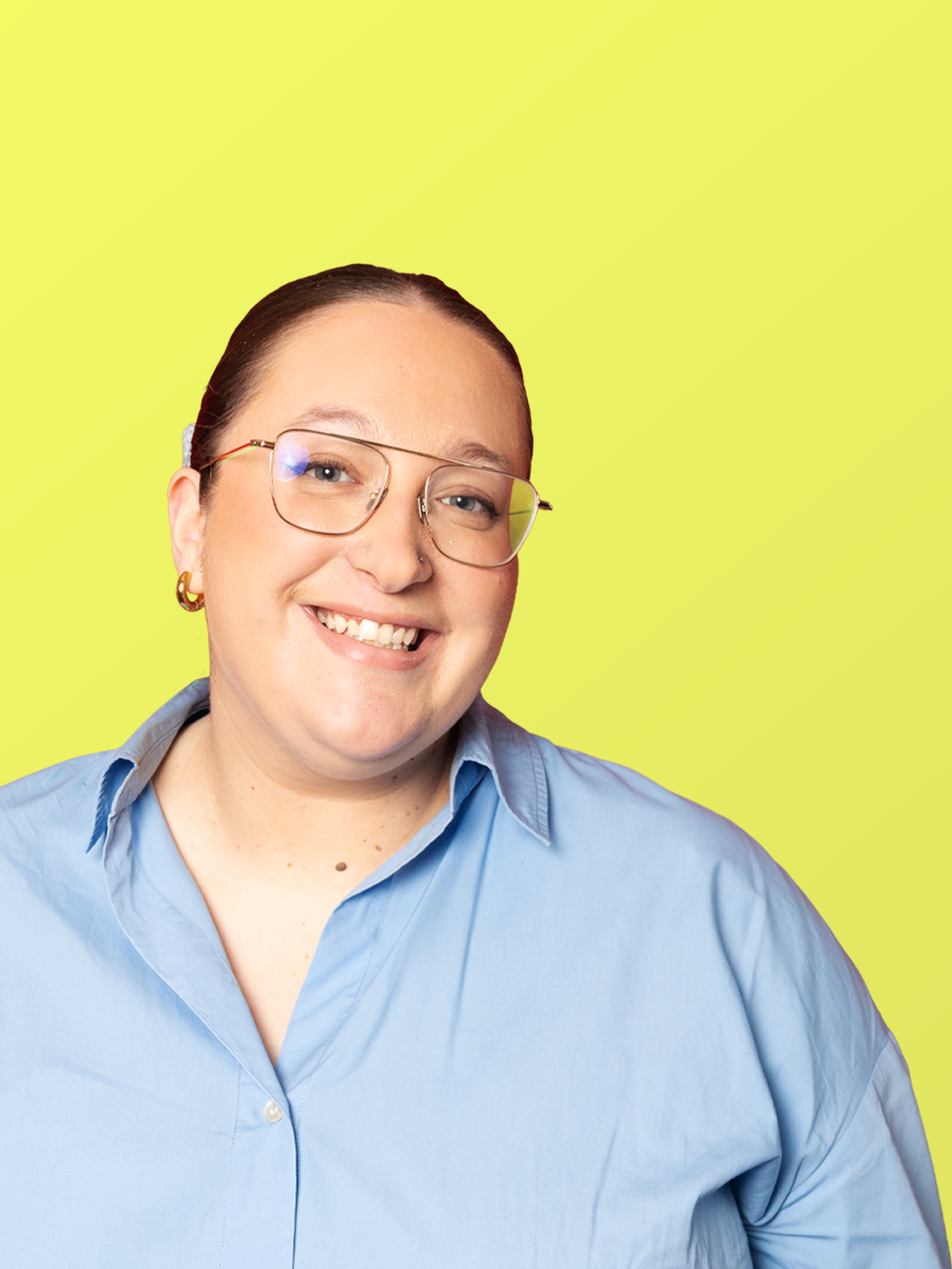 A smiling woman with short brown hair, wearing glasses, gold hoop earrings, and a light blue button-up shirt, stands against a bright yellow background.