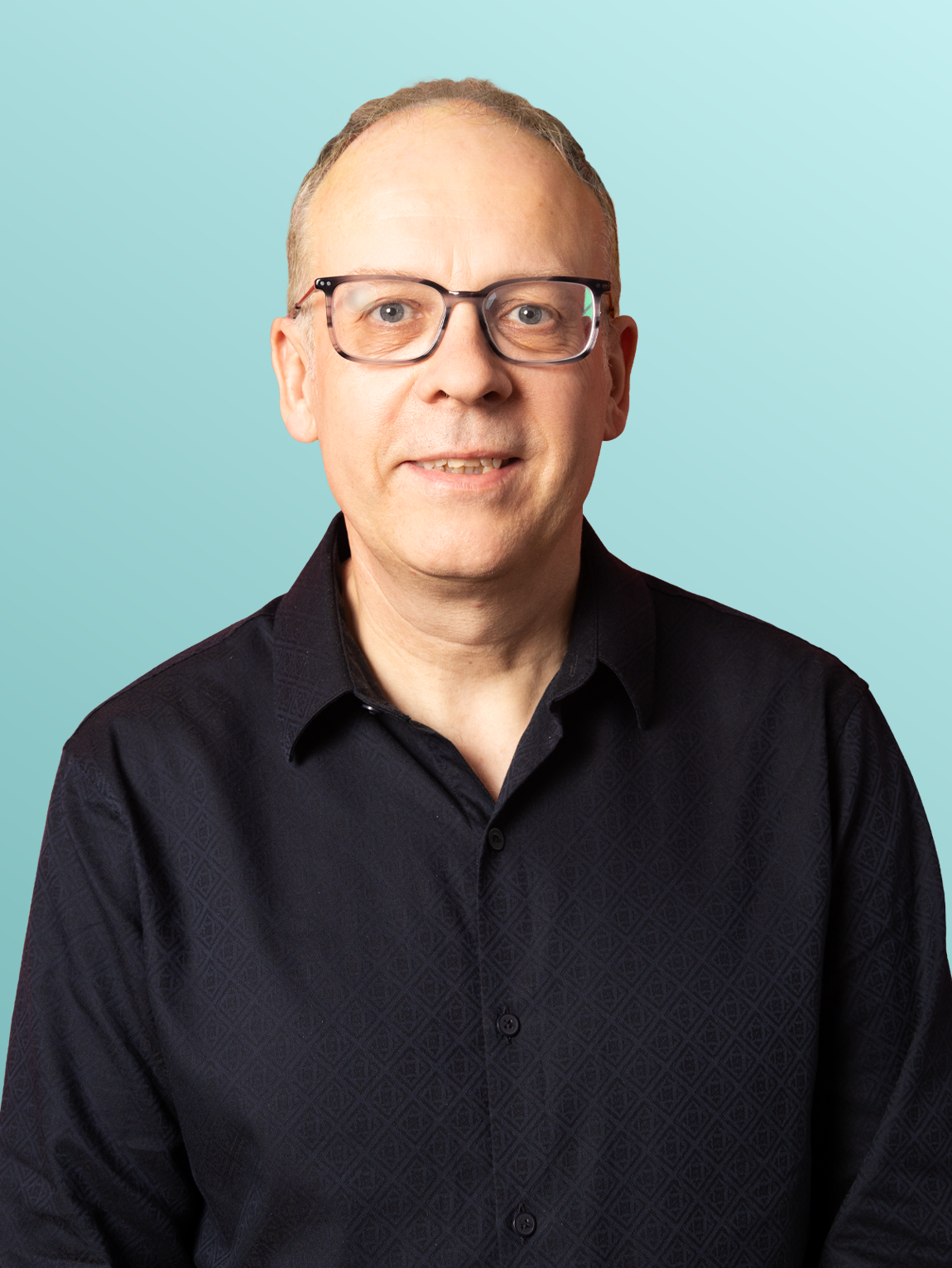 A man wearing glasses and a black button-up shirt stands against a light blue background.