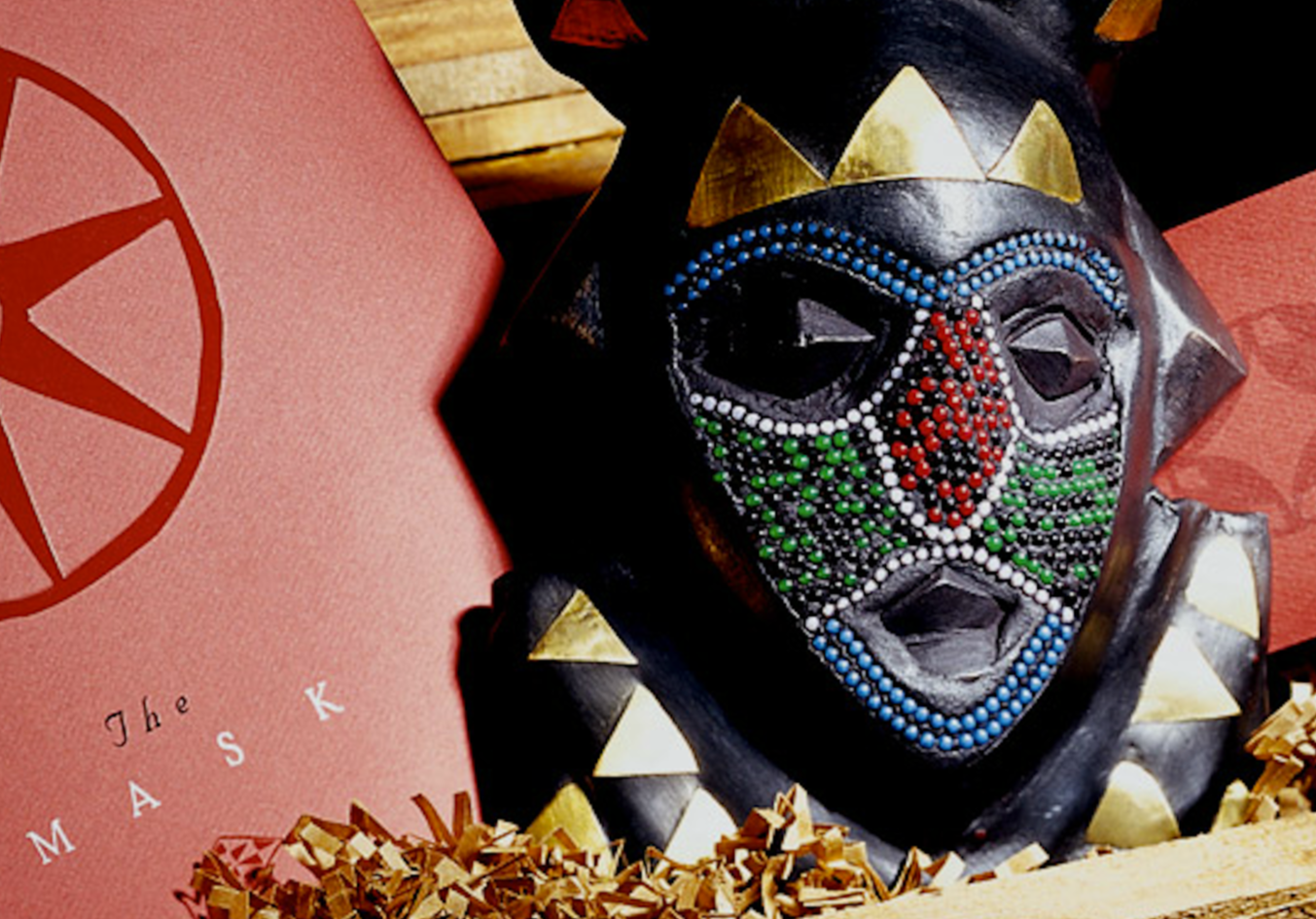 A decorative black tribal mask with colorful rhinestone embellishments and golden details, placed inside a wooden box with pink paper featuring the Masonic square and compass symbol.