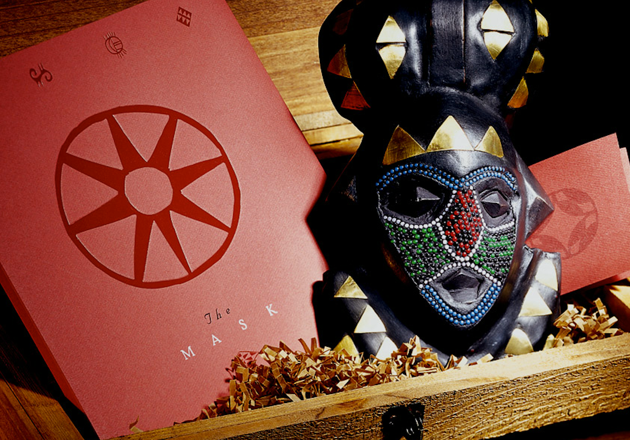 A decorative black tribal mask with colorful rhinestone embellishments and golden details, placed inside a wooden box with pink paper featuring the Masonic square and compass symbol.