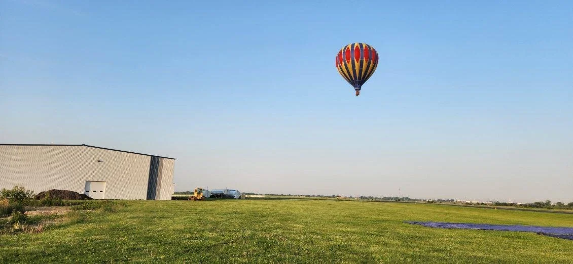A hot air balloon with red, yellow, and black stripes floating in the sky above a grassy field near a large industrial building.