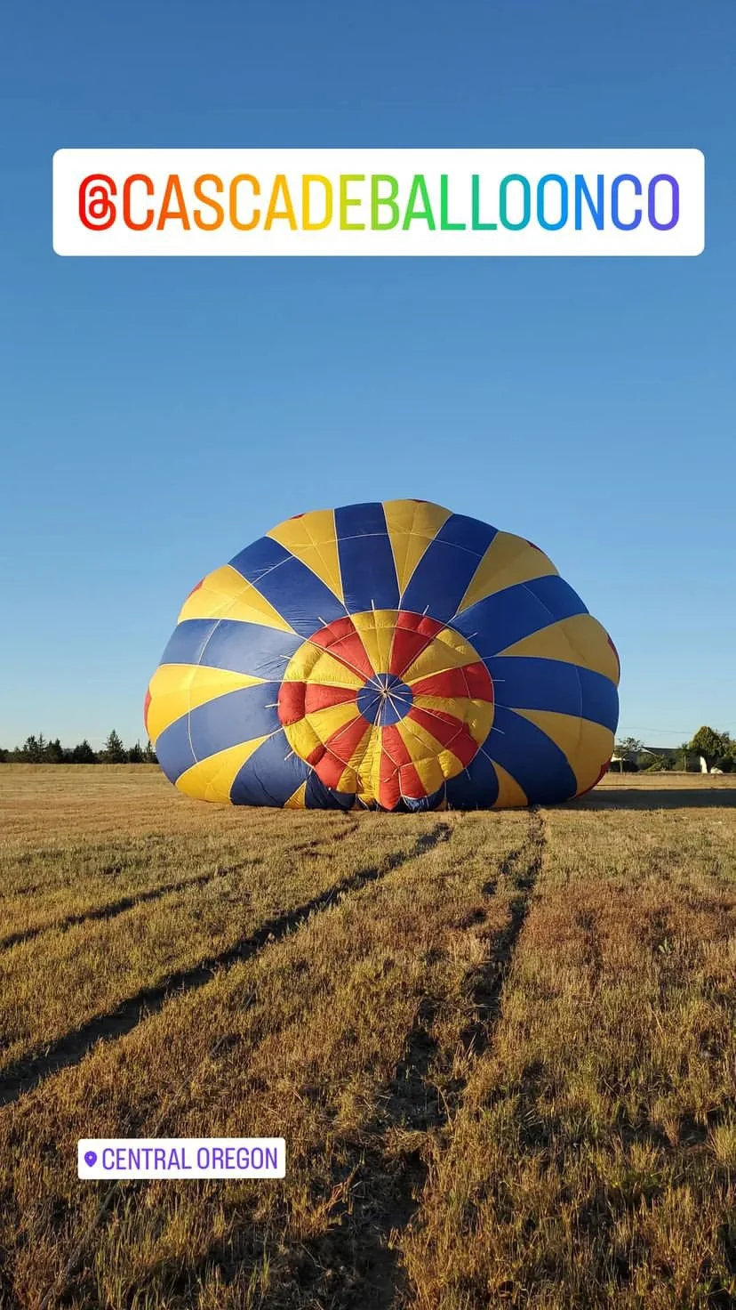 A colorful hot air balloon in the process of being inflated on a grassy field in Central Oregon, under a clear blue sky.