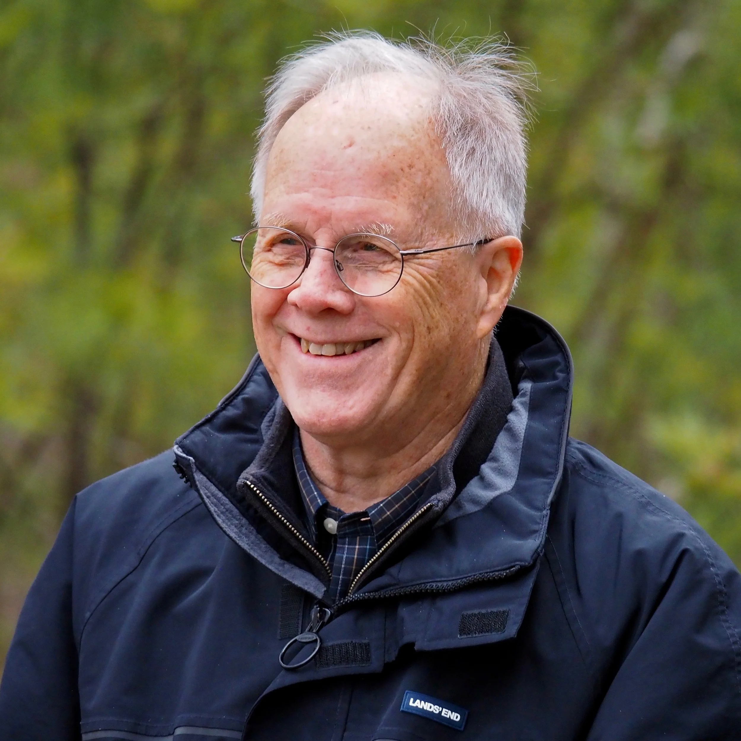A smiling elderly man with white hair and glasses, wearing a black jacket with a "LAND'S END" logo, outdoors with a blurred green foliage background.