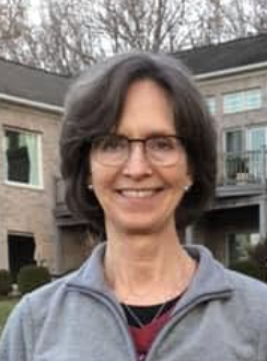 Portrait of a woman with short dark hair, glasses, smiling, wearing a gray zip-up jacket, outdoors in front of a house with a balcony and trees.