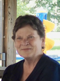 Portrait of a woman with short dark hair, smiling, wearing a dark top, background with colorful floatation devices and trees outside.