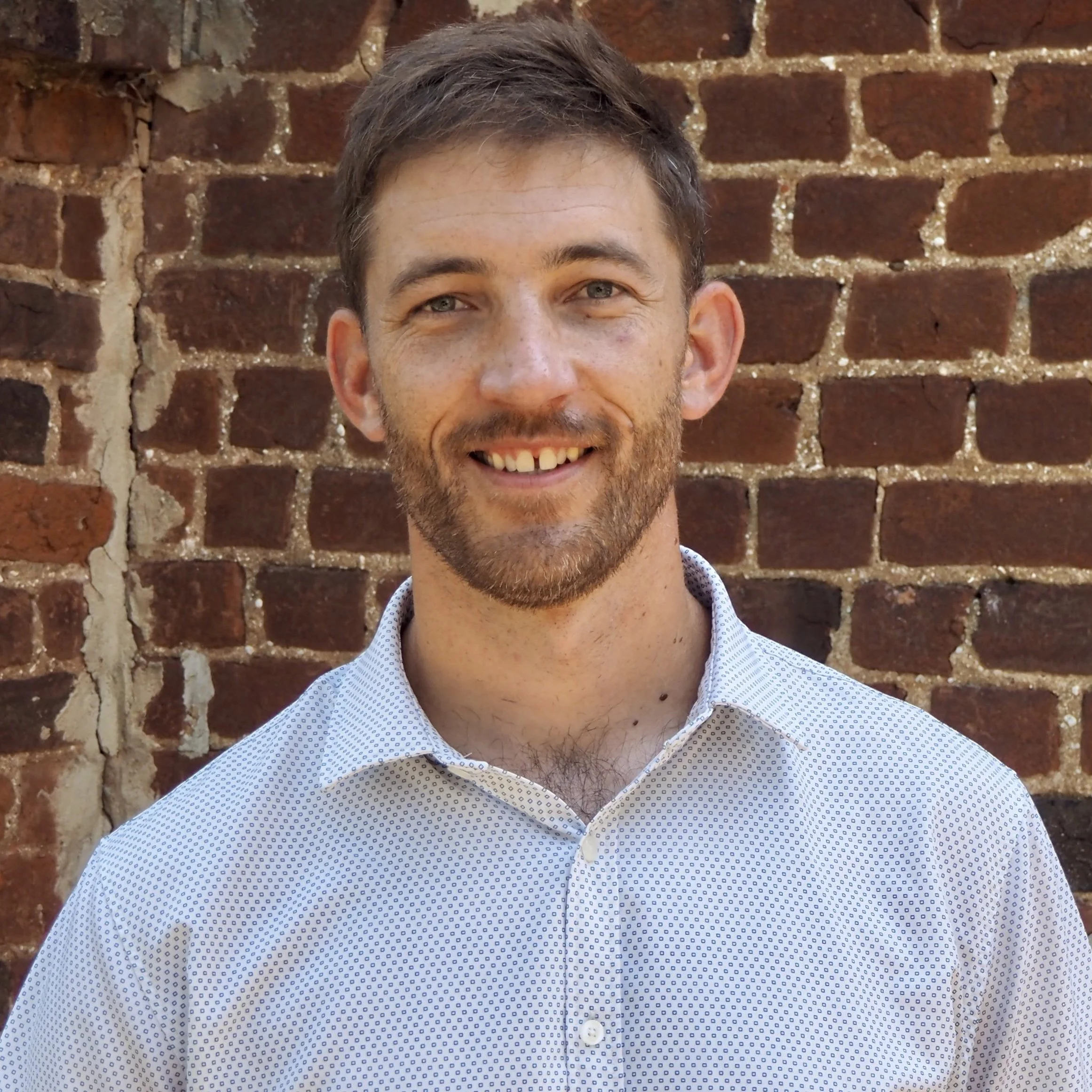 Close-up of a smiling man with light hair and a beard, wearing a light blue button-up shirt with small dark patterns, standing in front of a red brick wall.