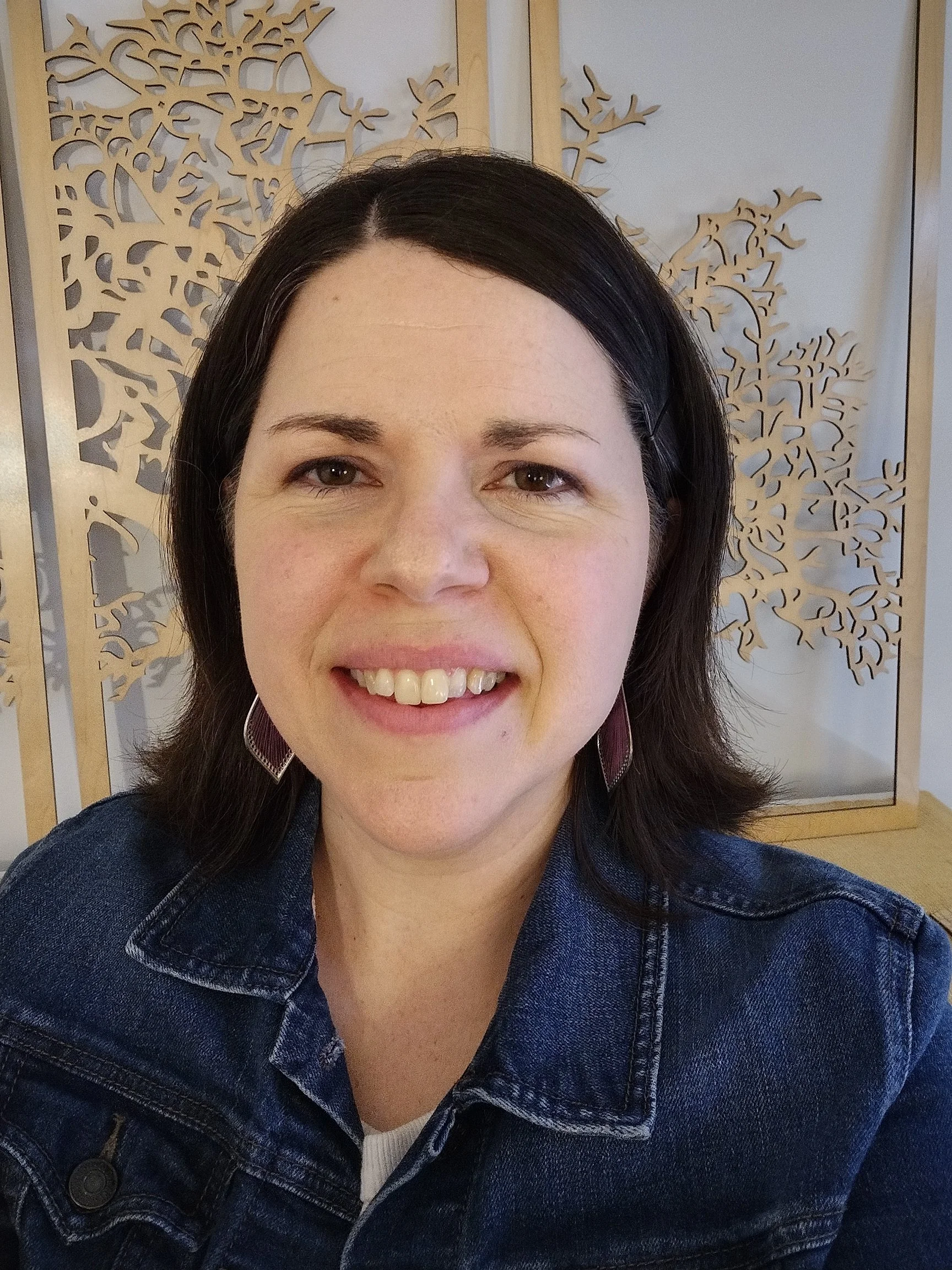 Close-up of woman with dark hair wearing a denim jacket, smiling with teeth showing, in front of decorative wooden wall panels with intricate cut-out design.