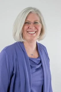 A smiling woman with shoulder-length gray hair wearing glasses and a blue blouse.