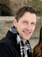 Smiling man with short brown hair and light skin, wearing a black coat and a black-and-white checkered scarf, outdoors with a stone wall and trees in the background.