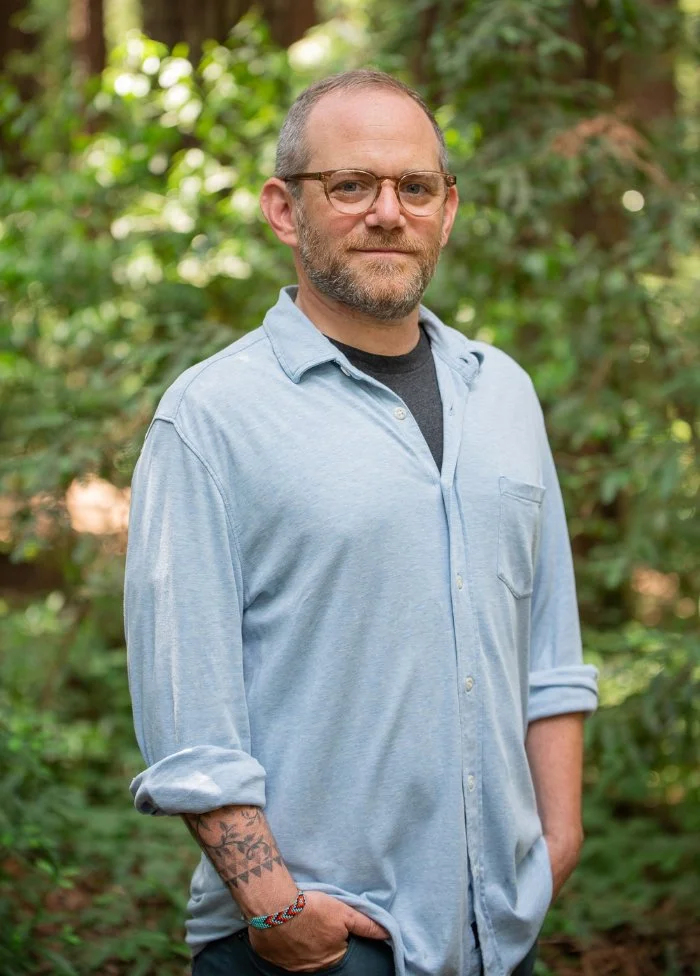 A man with glasses and a beard stands outdoors with greenery in the background, wearing a light blue button-up shirt with rolled-up sleeves and a black T-shirt underneath.