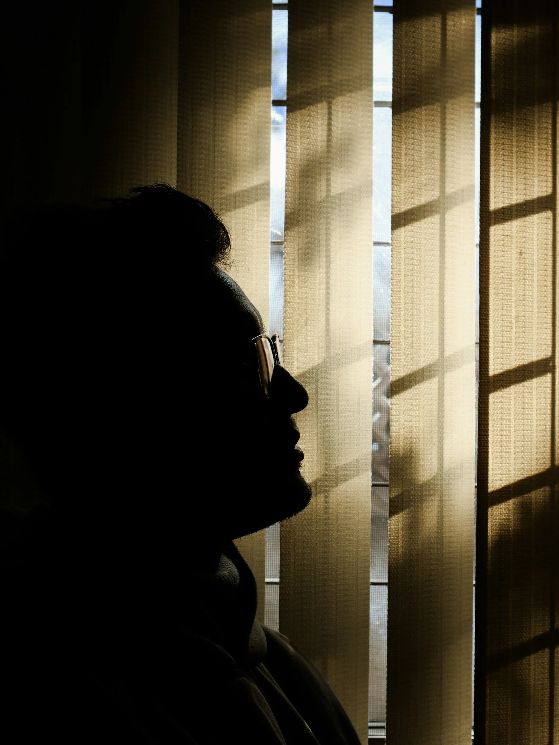 Silhouette of a person with glasses looking out a window with beige blinds, sunlight streaming through.