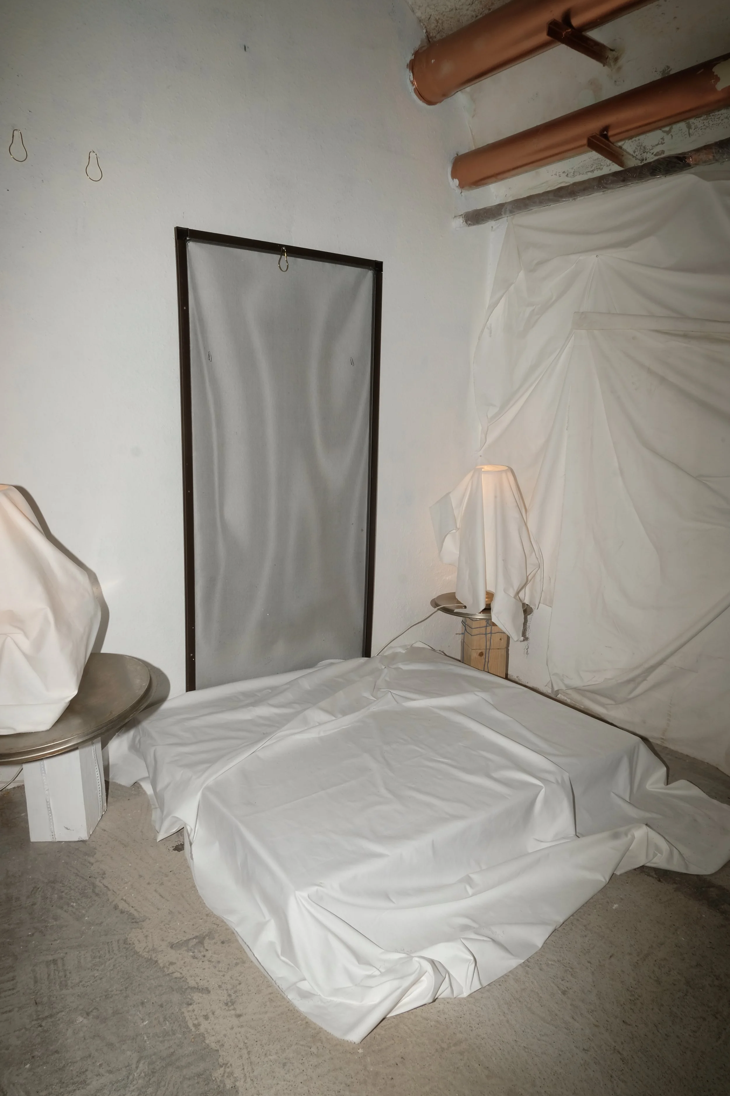 A room with a bed covered in white plastic, a small lamp with a white shade on a wooden block, a black powder-coated metal frame on the wall, and exposed copper pipes in the ceiling.