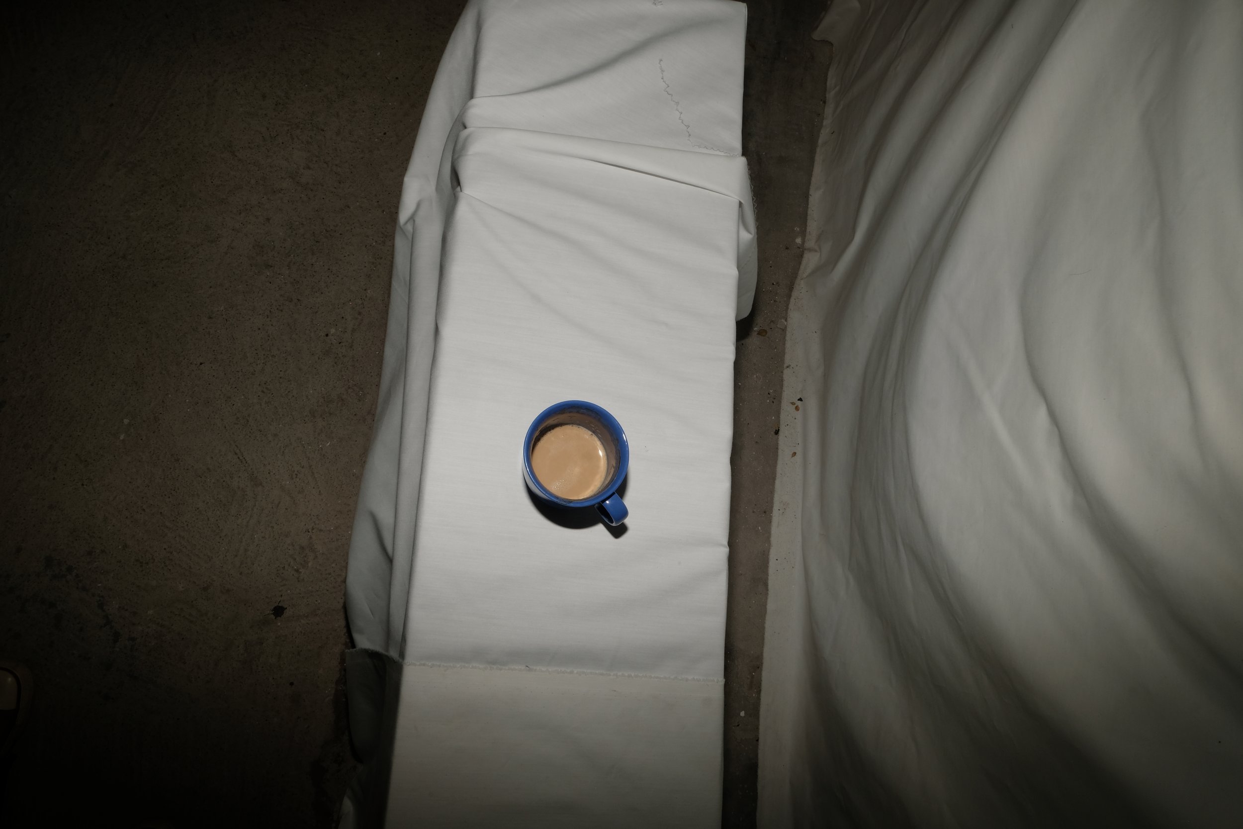Blue mug filled with coffee on a white bed. The bed has slight wrinkles in the white bedsheet, and there's a beige wall in the background.