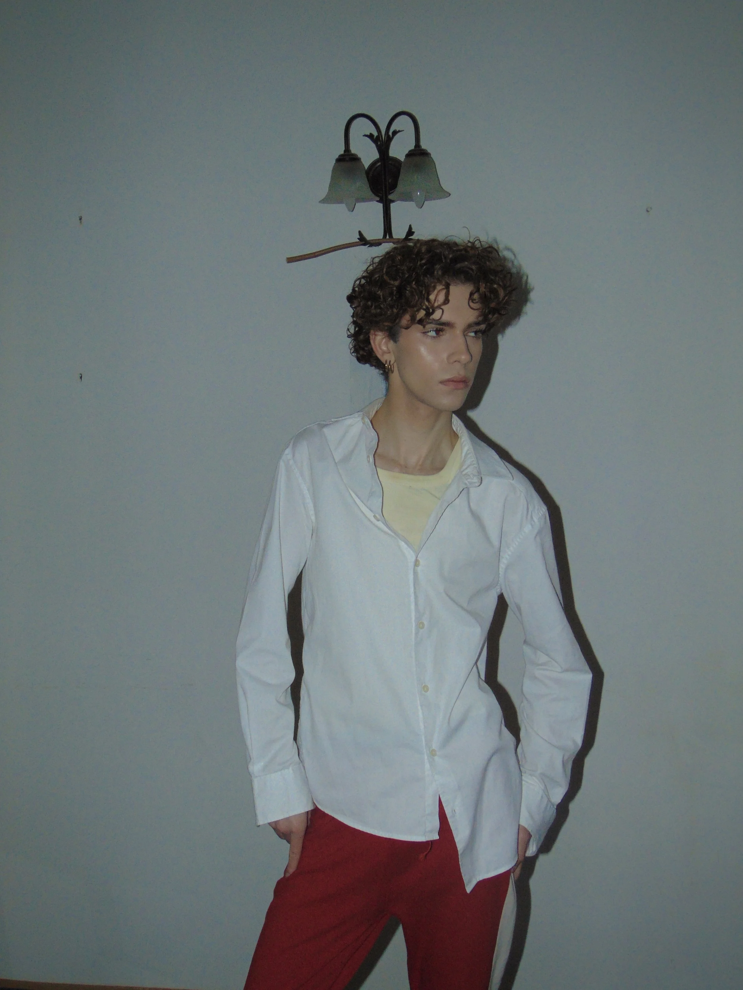Person with curly hair wearing a white shirt and red pants standing against a plain wall with a wall lamp above.