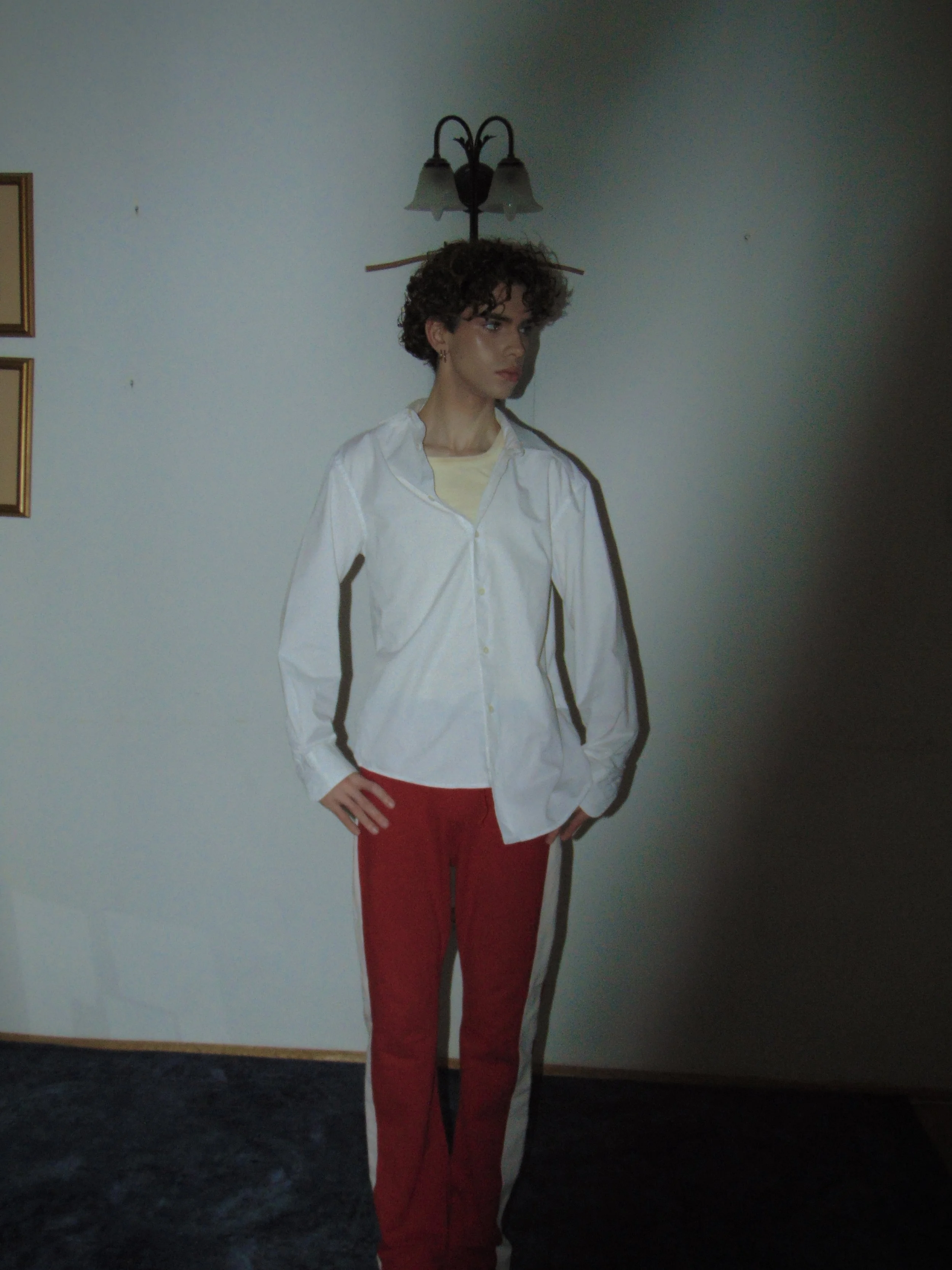 A woman with curly hair wearing a white shirt and red pants standing indoors against a plain wall with a lamp fixture above her head.