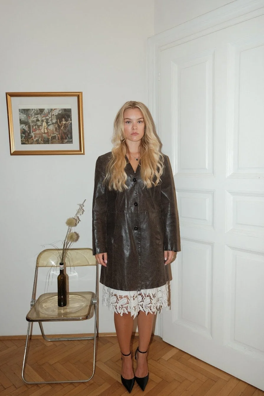 A young woman with long blonde hair standing indoors, wearing a long brown leather coat, lace dress, and black high heels, against a white wall with a framed picture and a chair with a vase of dried flowers nearby.
