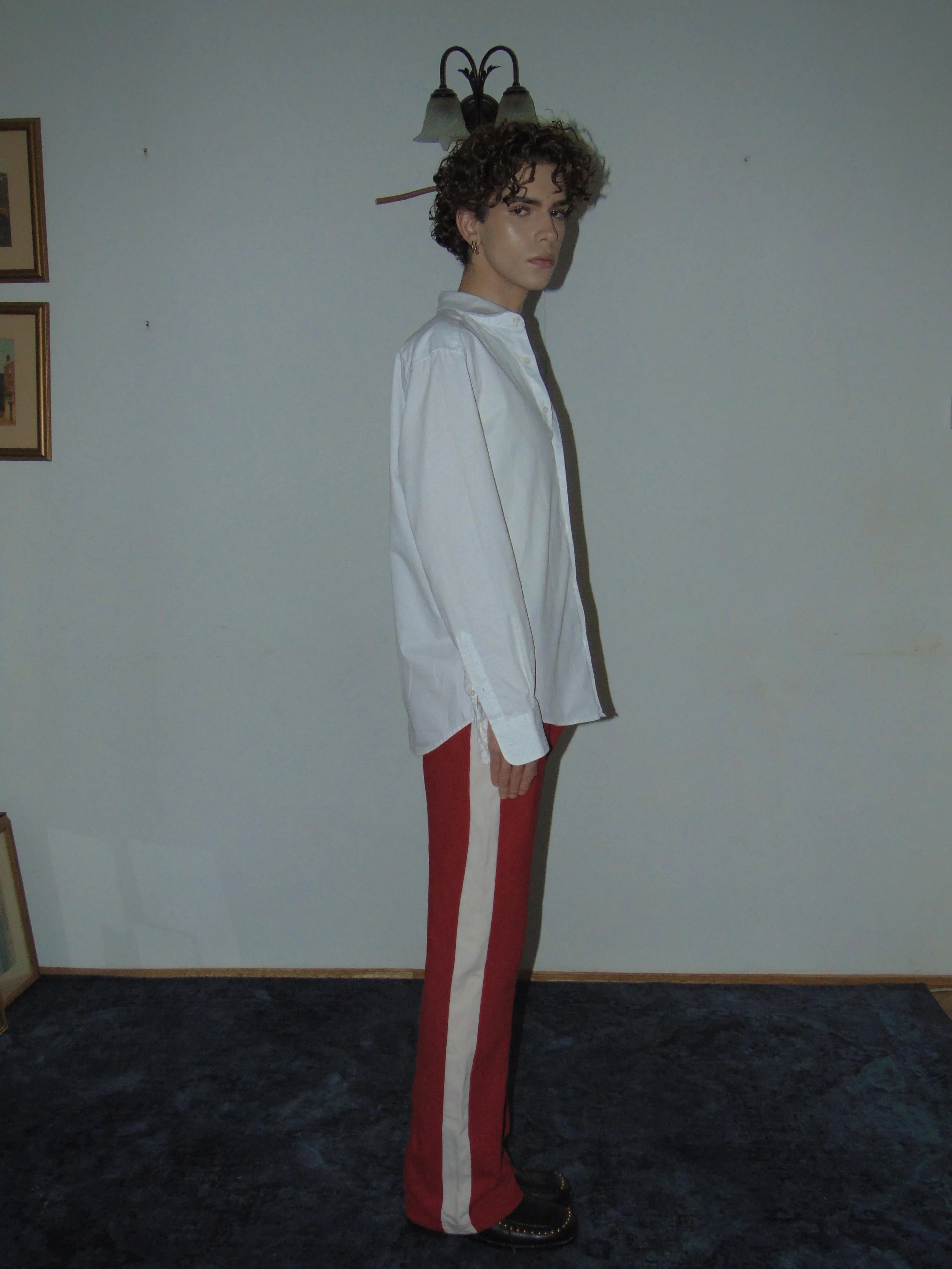 Person with curly hair wearing a white shirt and red pants with white stripe, standing in room with white wall and picture frames.