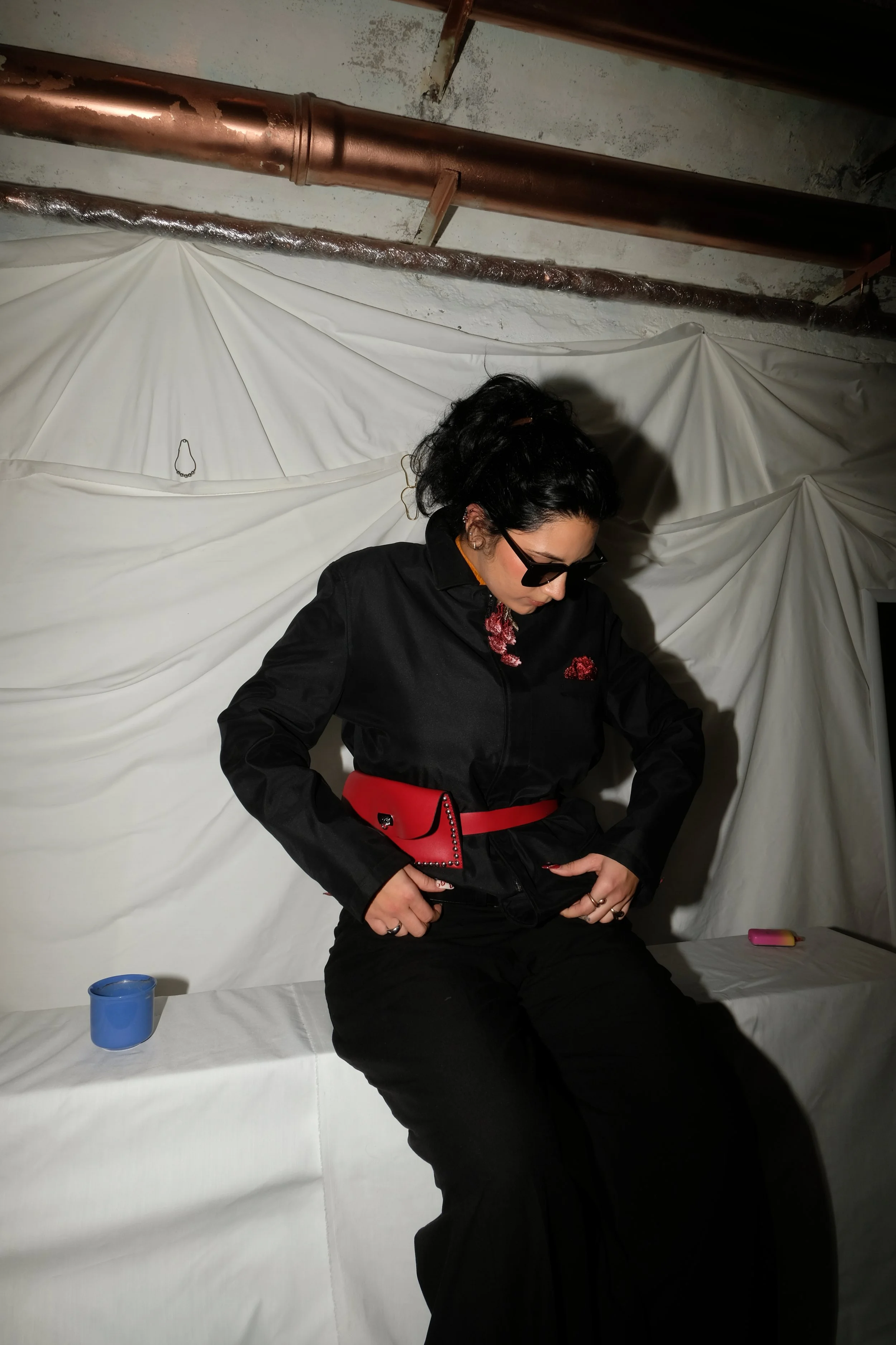 A woman with dark curly hair, wearing sunglasses, a black jacket, and black pants, sitting on a white surface with a white draped backdrop in the background. She has a red belt and matching red purse, with small objects placed on the surface beside h