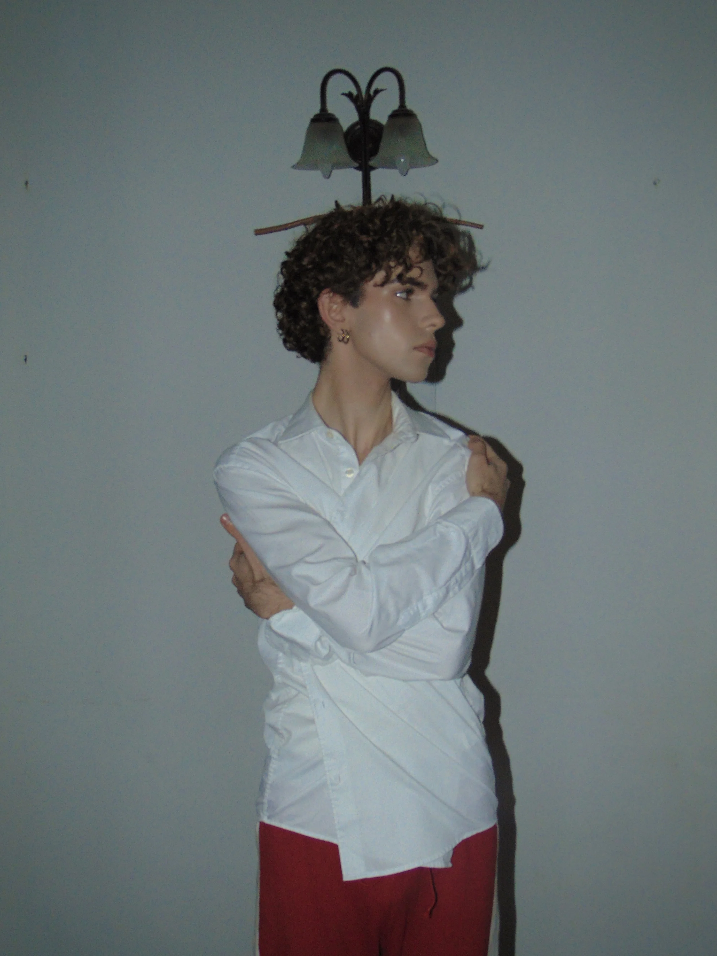 A woman with short curly hair and earrings stands against a plain wall, with a wall-mounted light fixture casting a shadow behind her.