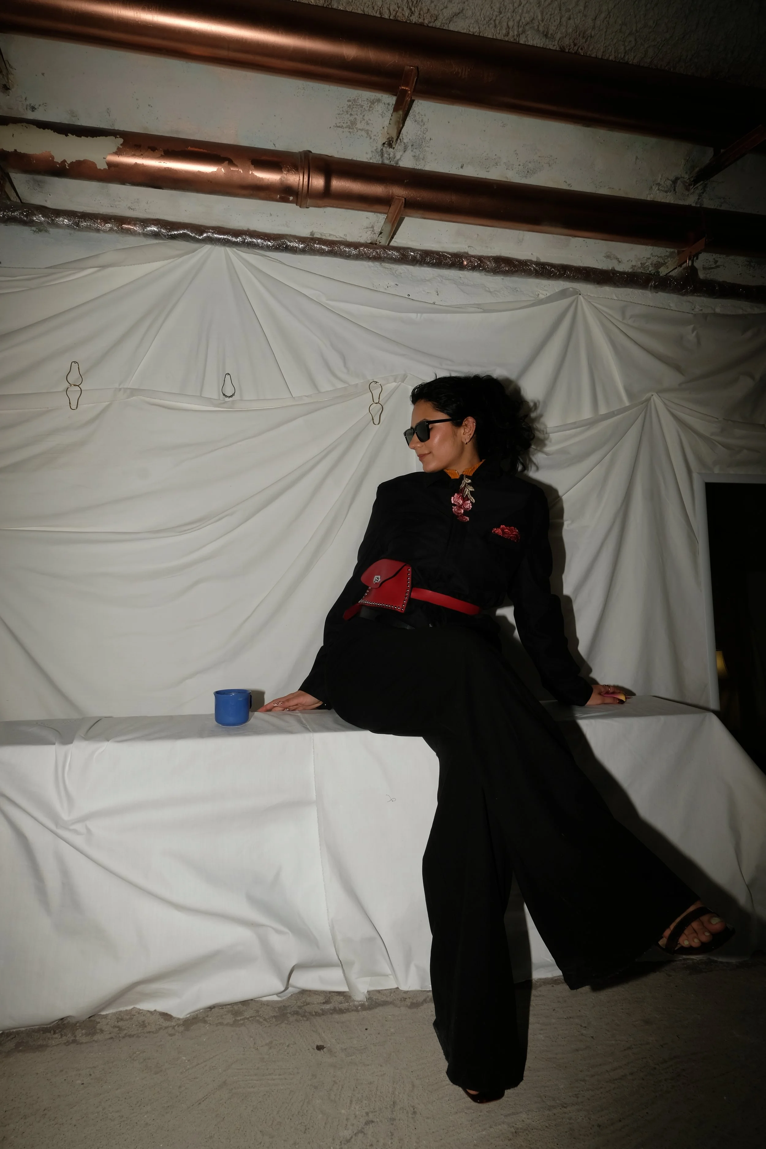 A woman dressed in black with a red belt and purse, black sunglasses, and black sandals, sitting on a white surface against a backdrop of draped white fabric with metal hooks, in an industrial or unfinished space with exposed pipes and concrete floor