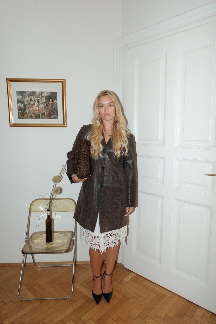 A young woman with long blonde hair standing indoors near a closed white door. She is wearing a black leather jacket, a white lace skirt, and black high-heeled shoes. She is holding a brown textured clutch bag. To her left, there is a small transpare