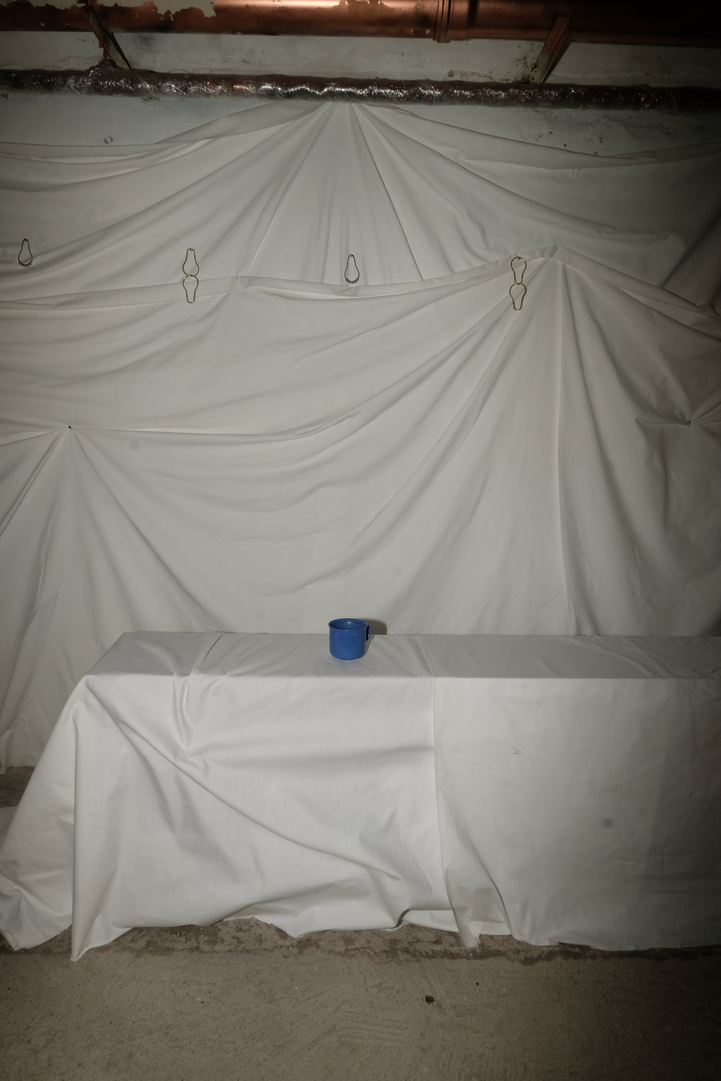 A white draped fabric backdrop with metal hooks, a table covered with white cloth, and a small blue cup on the table. The scene appears to be inside a basement or storage area with exposed pipes and concrete floor.