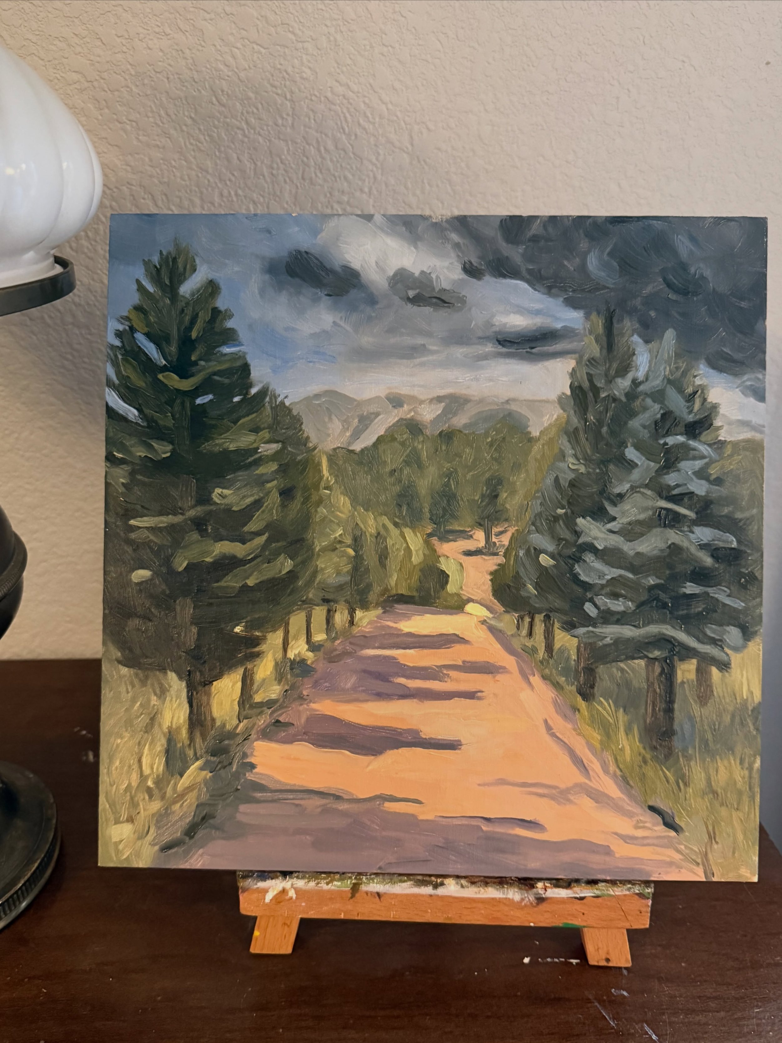 Oil painting of a dirt road through evergreens toward mountains under a storm sky in Woodland Park, Colorado.