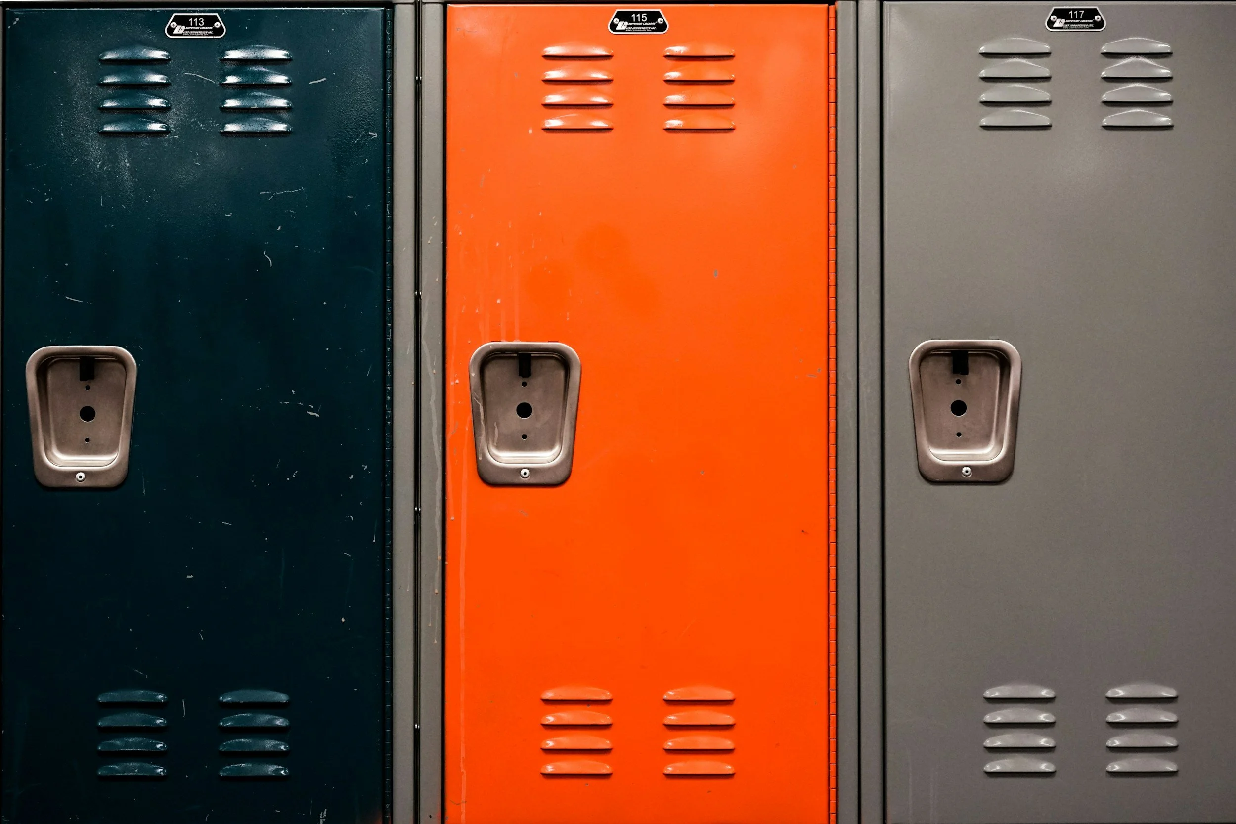 Three school lockers in dark green, orange, and gray colors, with ventilation slits and unlocked handles.