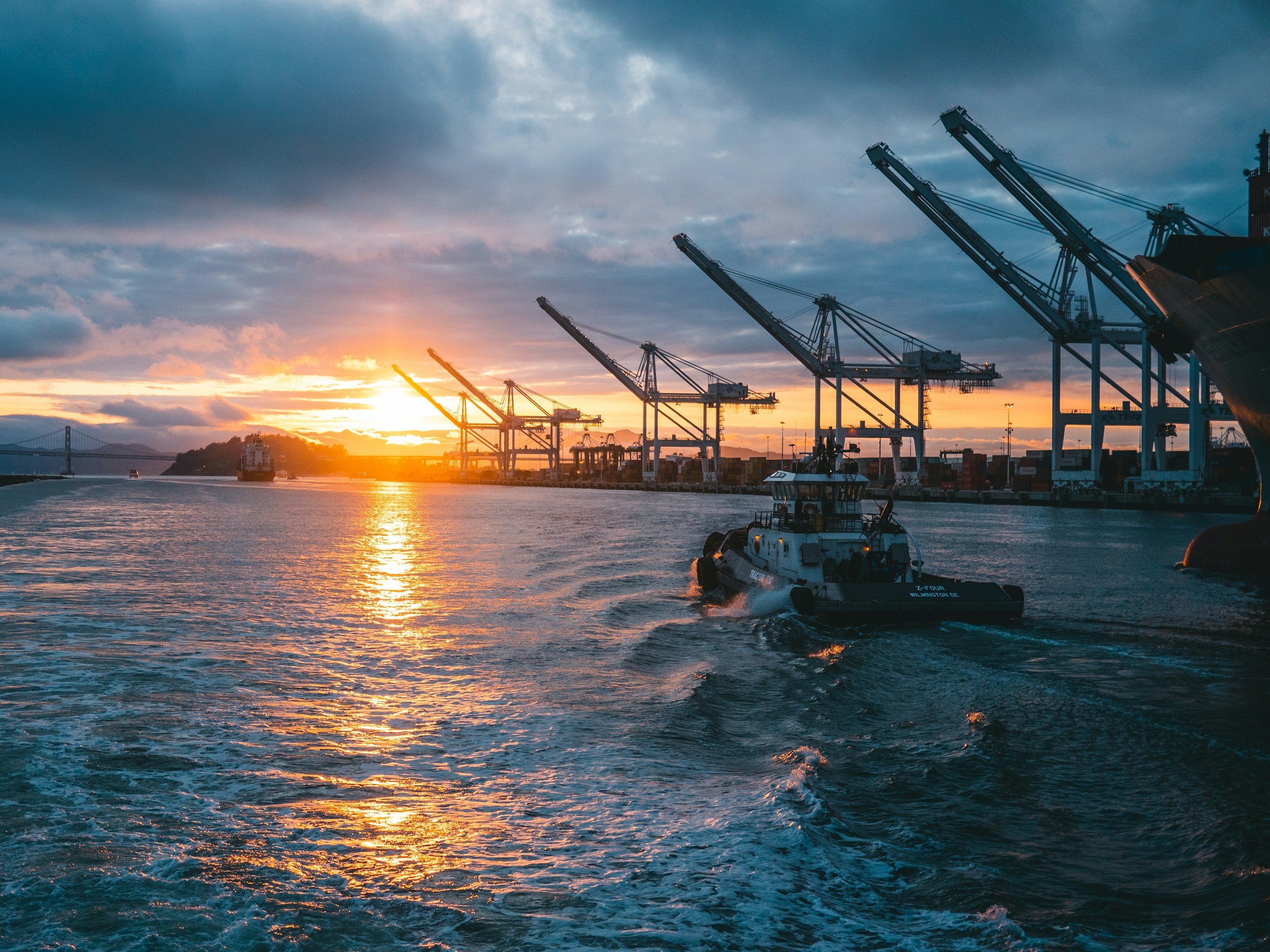 View of a harbor with boats and cargo cranes at sunset, with a tugboat in the foreground and ships near the port.