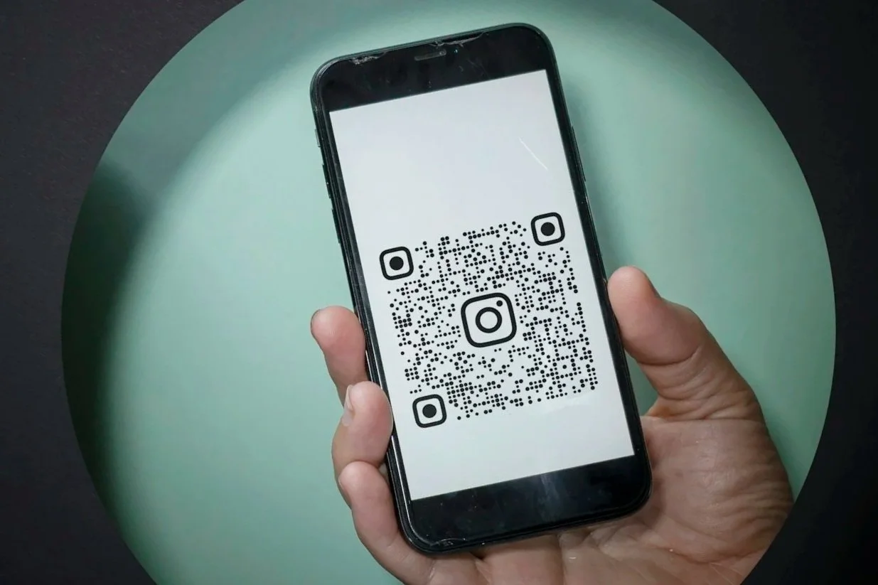 A person holding a smartphone displaying a QR code that has the Instagram logo in the center and corners, set against a dual-colored background with a greenish circular area.