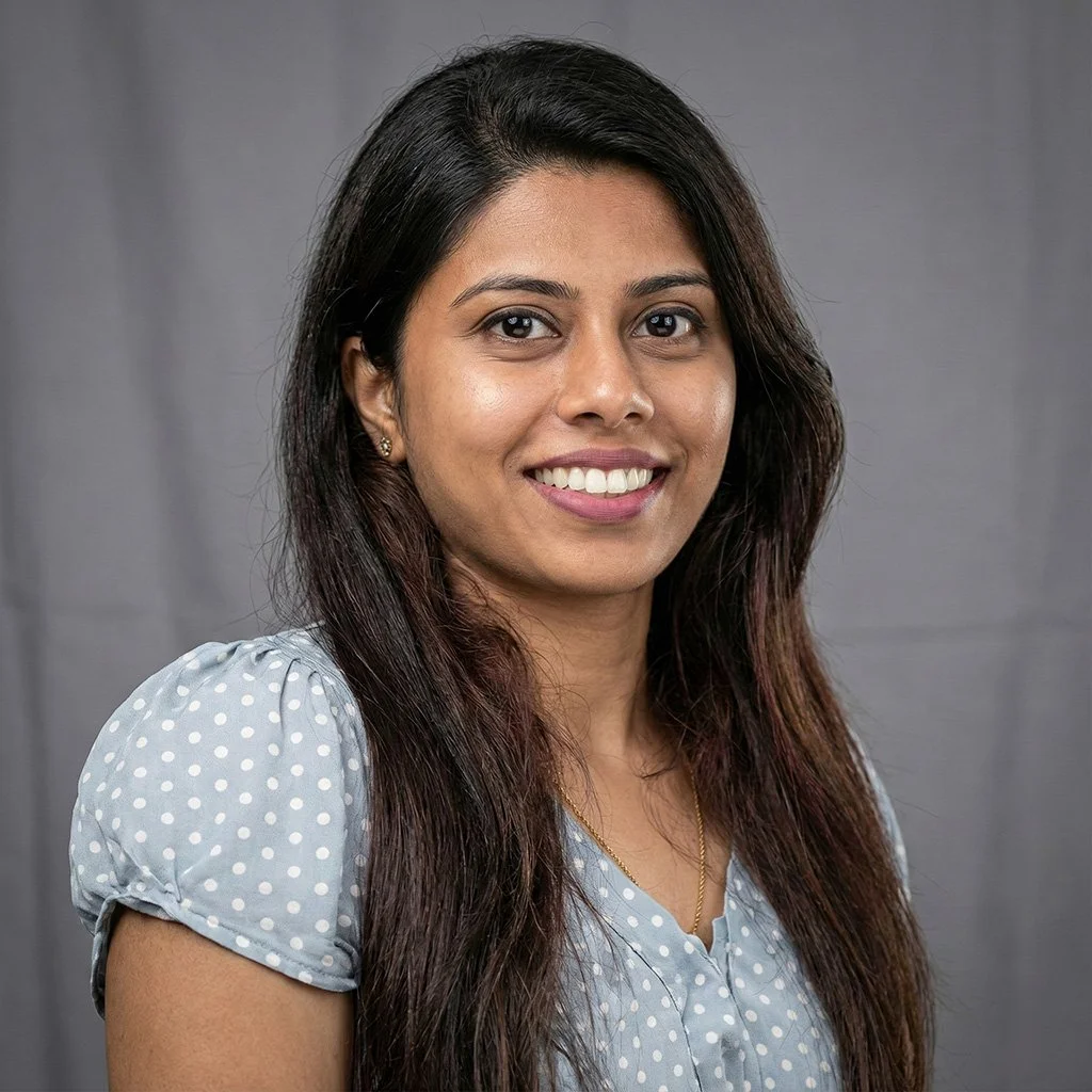 A woman with long dark hair wearing a light blue polka dot top, smiling in front of a plain gray background.