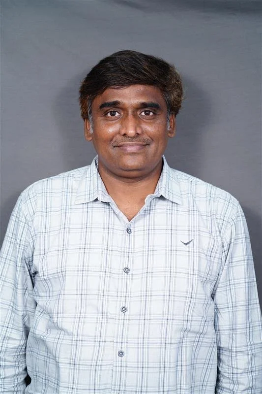 A man with short black hair and medium brown skin, wearing a light-colored plaid button-up shirt, standing against a gray background.
