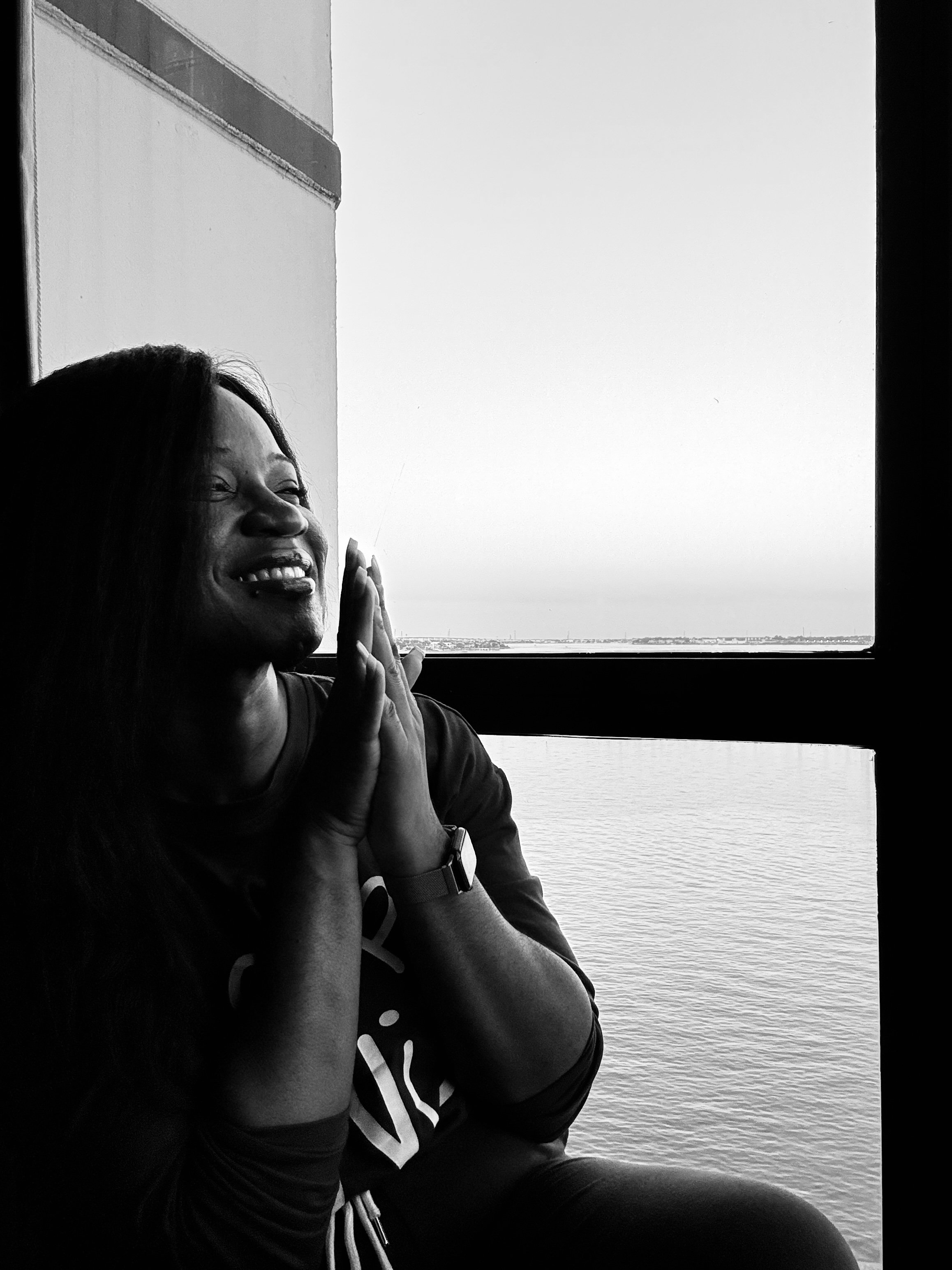 A woman with long dark hair smiling and holding her hands together near her face, sitting indoors near a large window overlooking water and a distant shoreline.