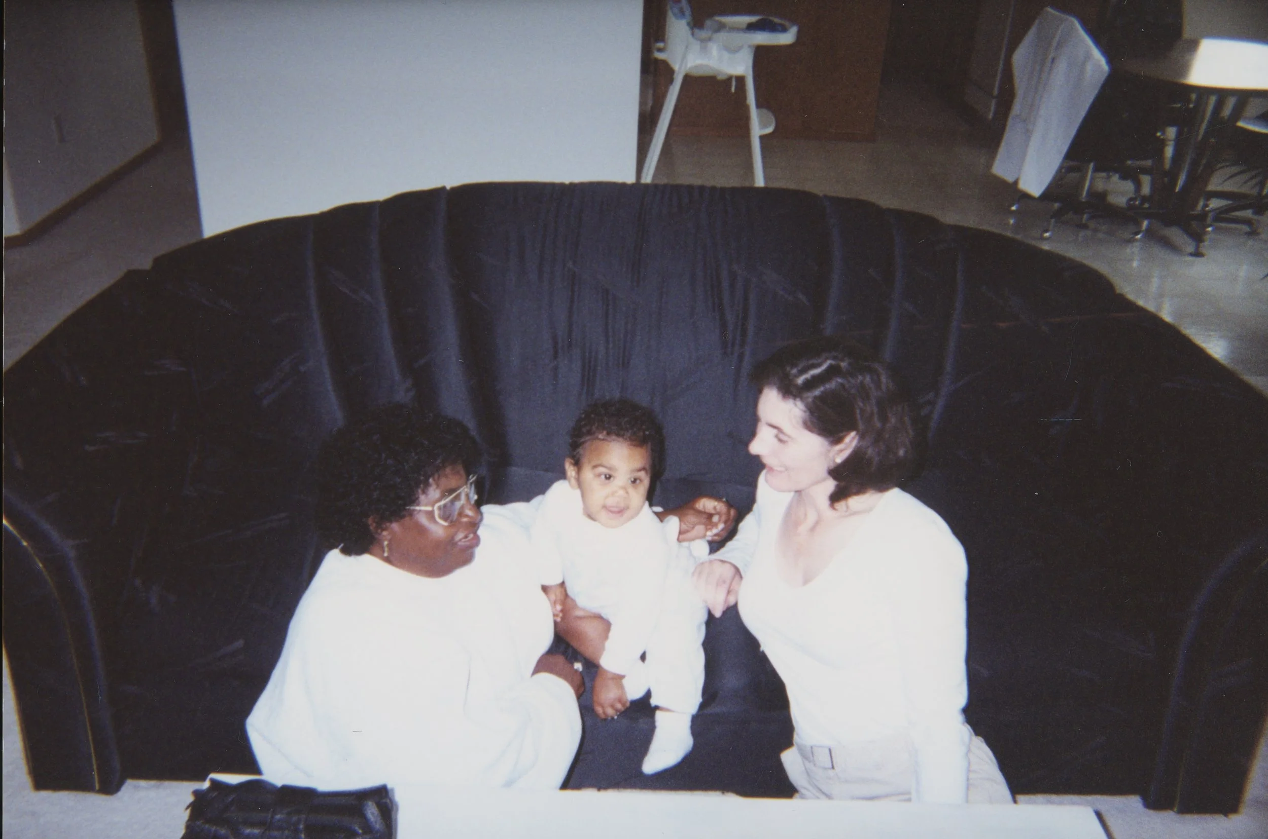 A woman, a child, and another woman sitting on a dark blue velvet couch in a living room, with the child in the middle holding hands with both women.