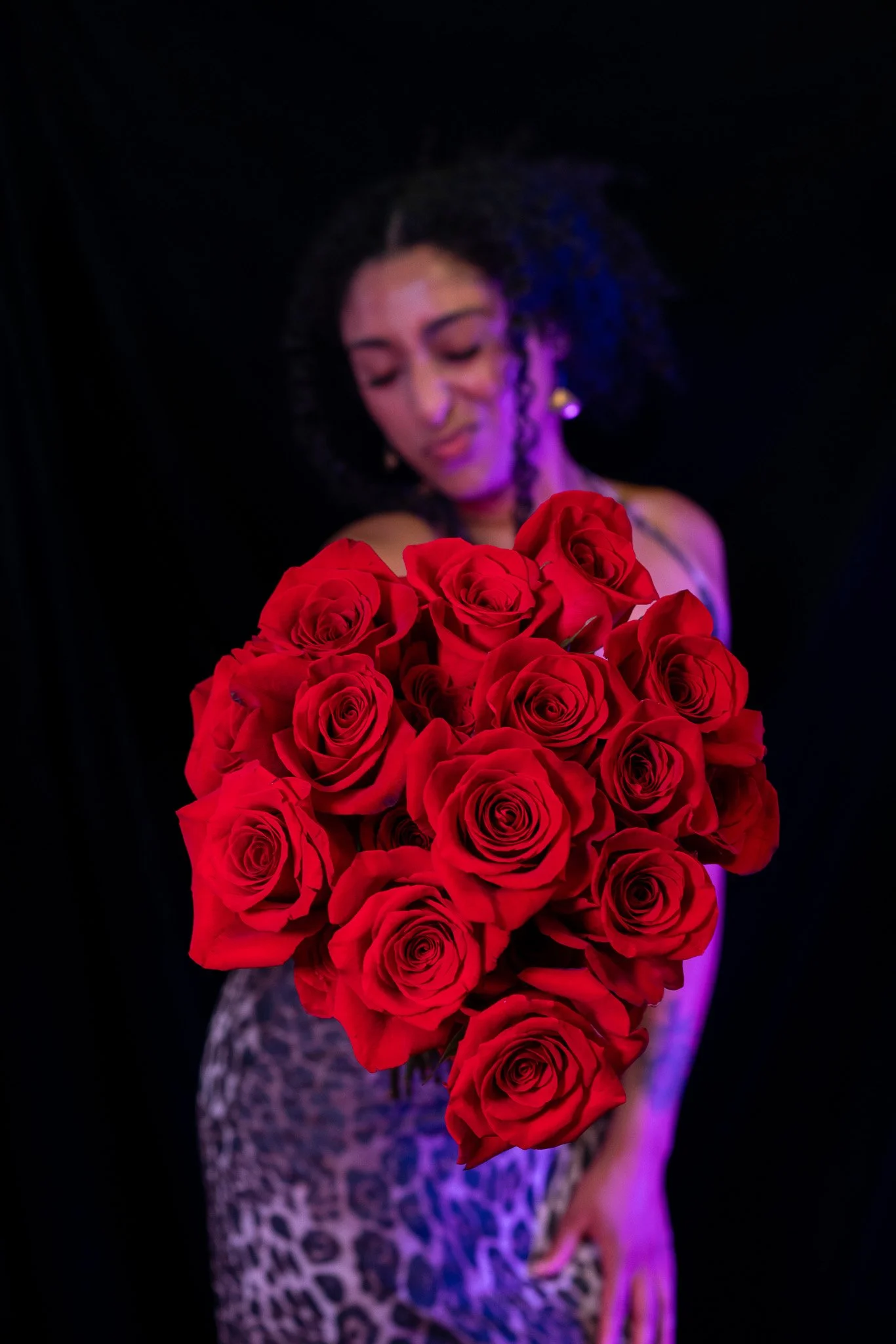 A woman holding a bouquet of red roses, with a blurred background and purple lighting.