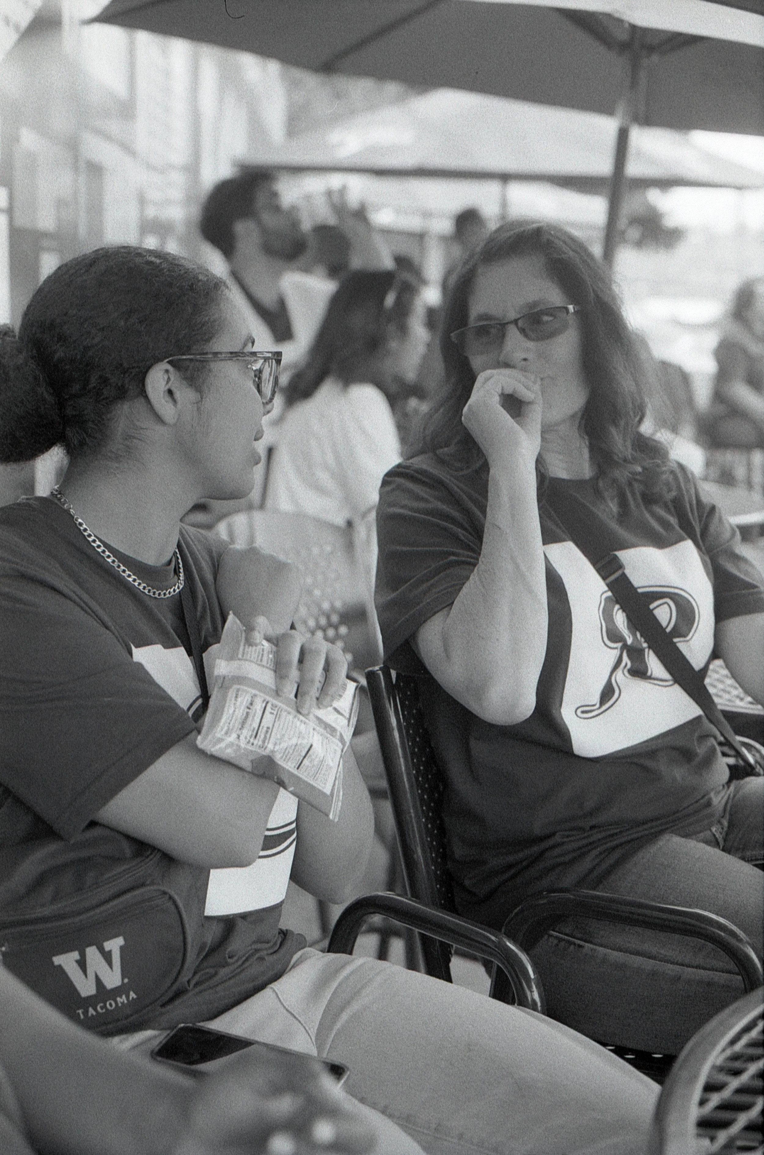 Two women sitting outdoors, engaged in conversation. The woman on the left has glasses, a chain necklace, holding a snack. The woman on the right has glasses, wavy hair, and with her hand near her mouth. In the background, there's a man drinking from a cup and other people sitting under umbrellas.