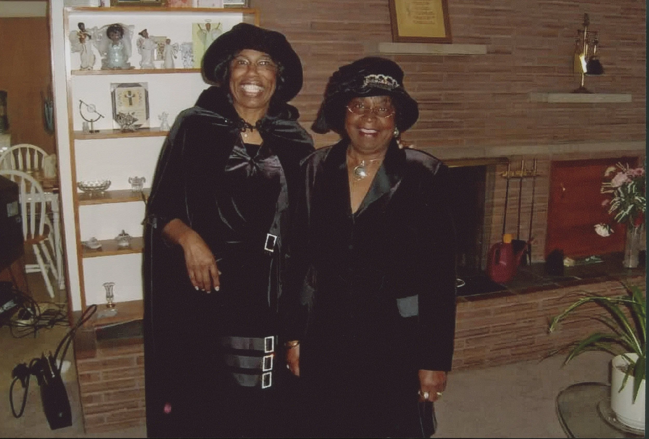Two women in black velvet dresses and hats standing next to each other indoors in front of a brick fireplace and wooden wall, smiling at the camera.